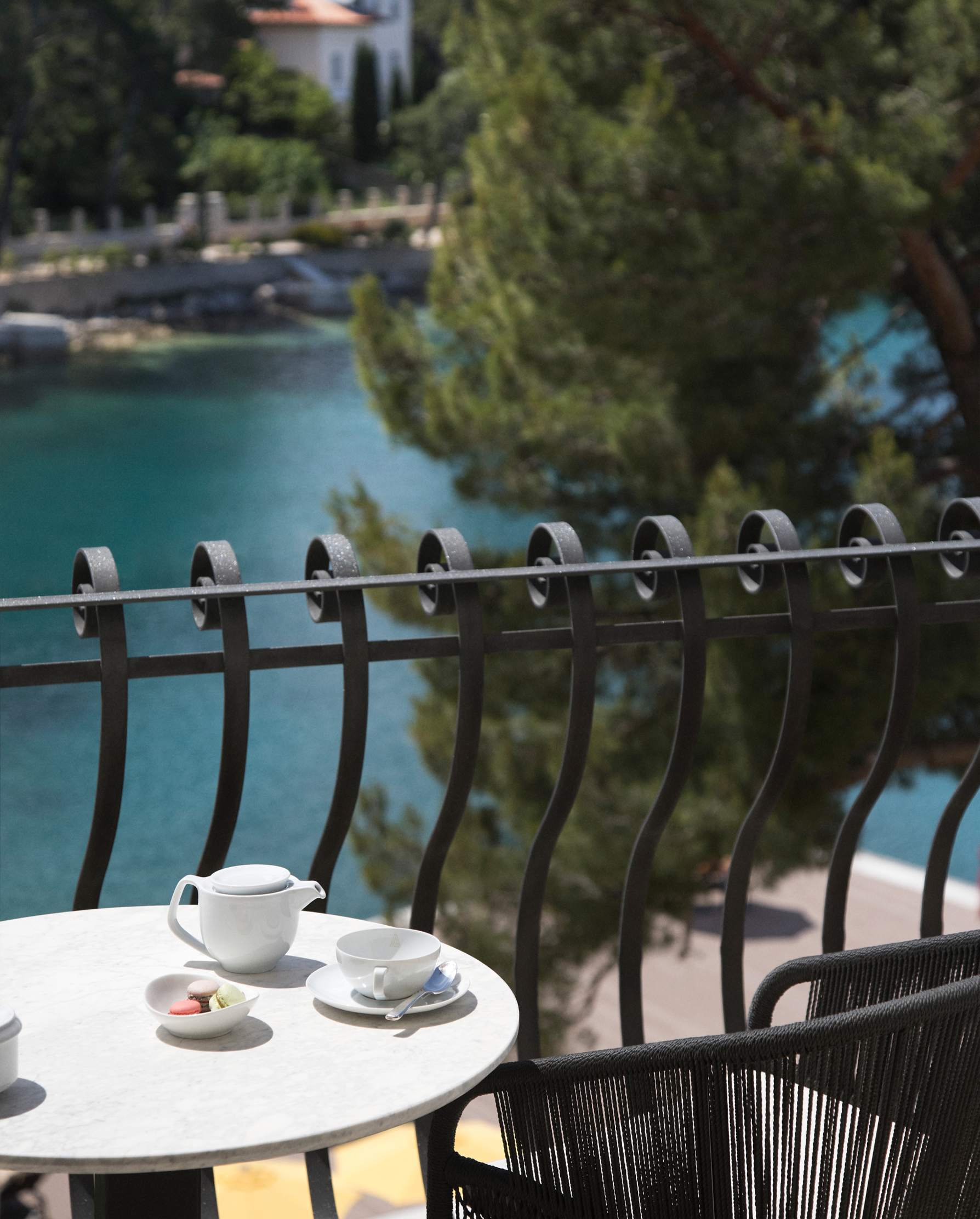 Two cups of coffee on a balcony table with a view of a serene blue sea and lush greenery.