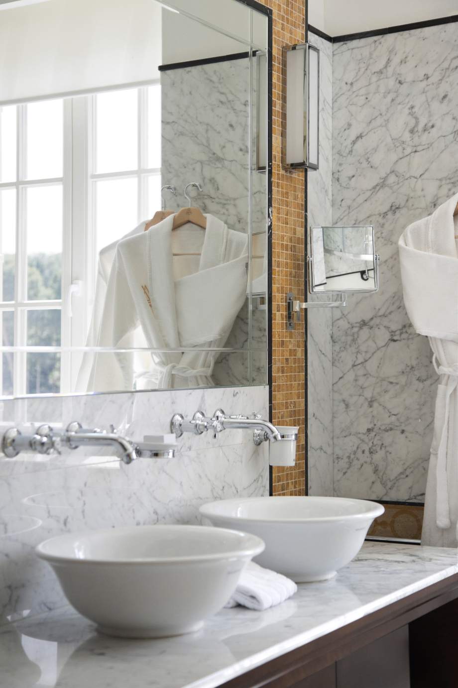 A luxurious bathroom with marble walls, twin vessel sinks, and a bathrobe hanging on a hanger with a window in the background