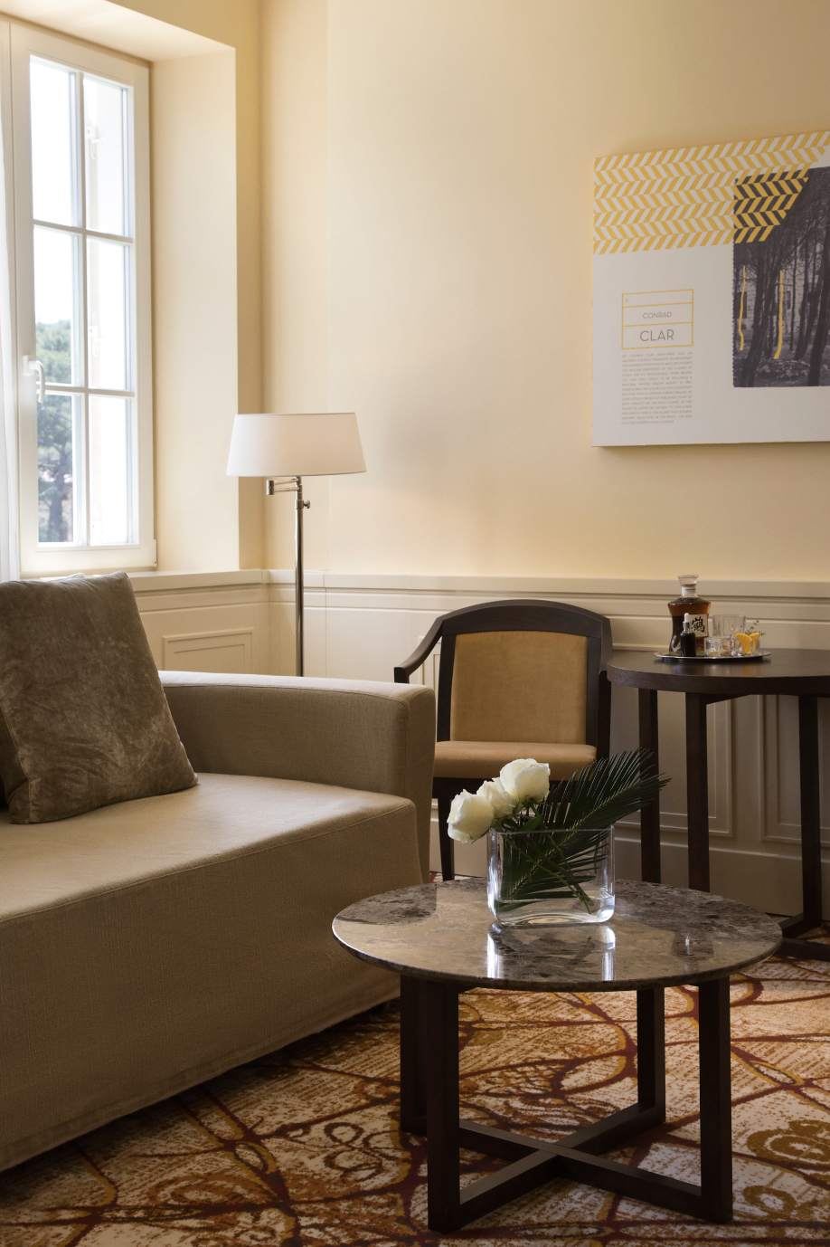 A cozy corner with a beige sofa, a round marble coffee table with flowers, beside a wooden chair and a small table under an elegant wall lamp