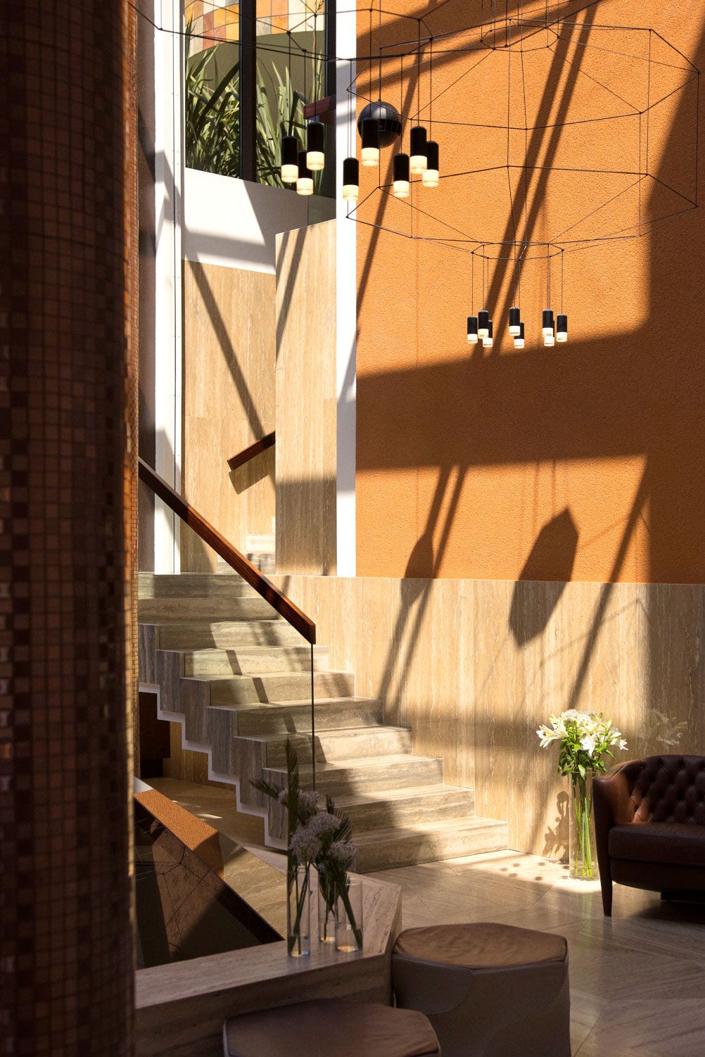 A modern atrium with natural light casting shadows on the warm-toned walls, featuring a staircase, hanging pendant lights, and a cozy seating area