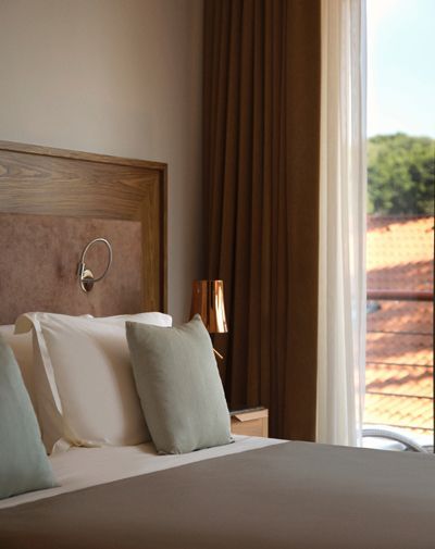 Comfortable bed with soft pillows beside a window with draped curtains, with terracotta rooftops visible outside