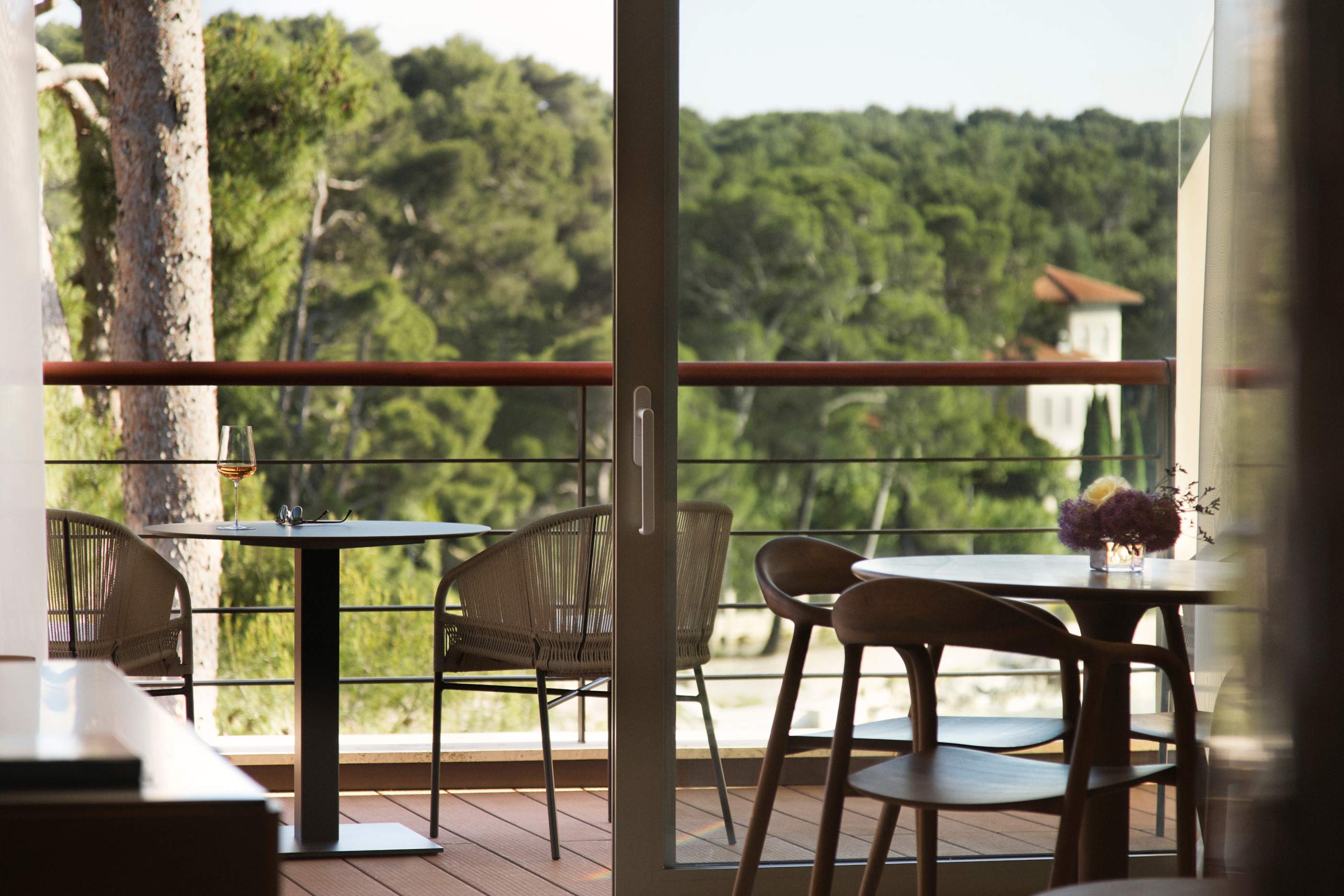 View from inside looking out onto a balcony with wicker chairs and a table set against a backdrop of a forested landscape