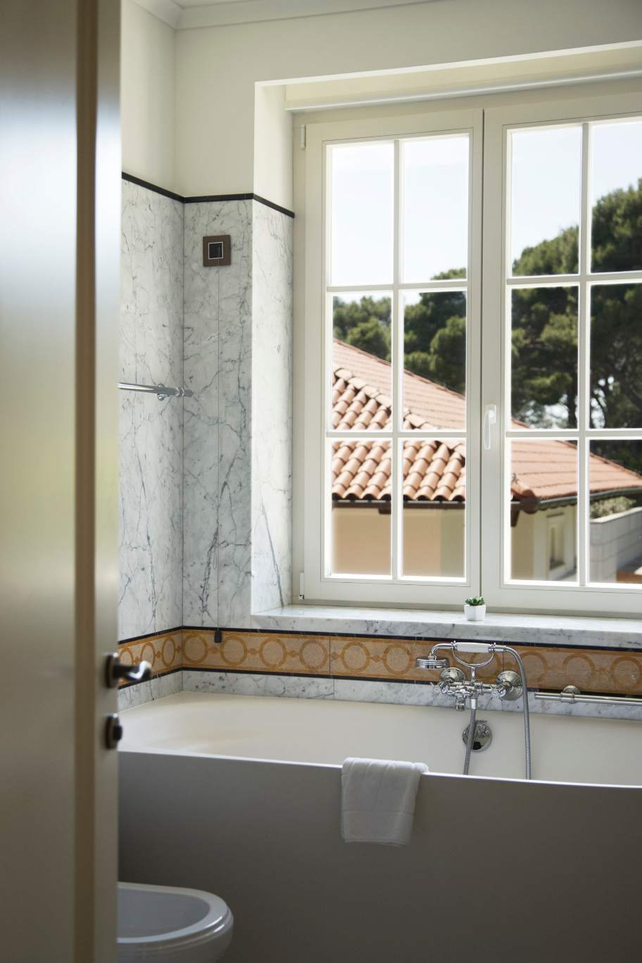 A bathroom featuring a tub with marble surround, accented with decorative tiles, and a window offering a view of trees and rooftops