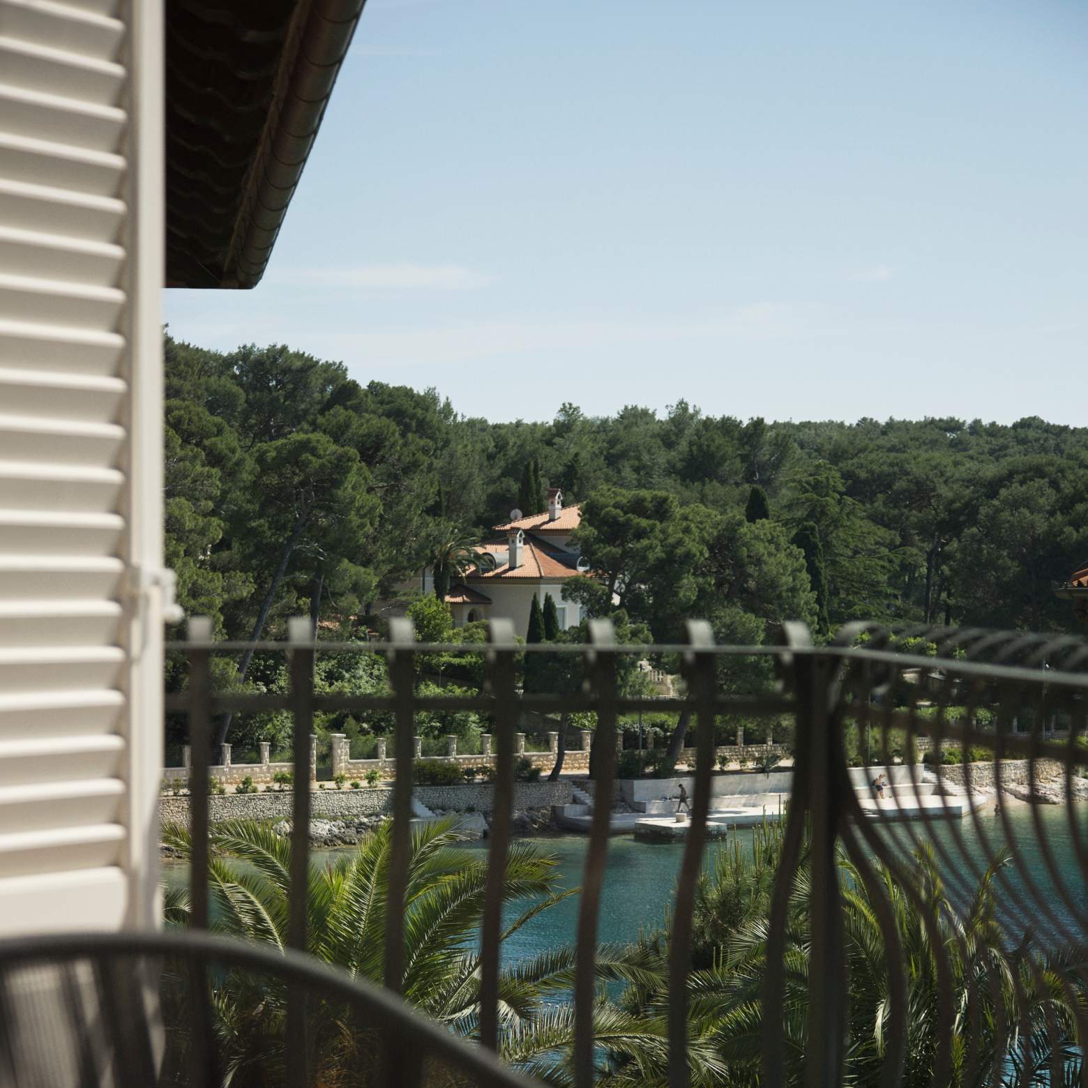 A balcony view framed by white shutters, overlooking a serene bay and lush greenery, with a glimpse of a coastal promenade