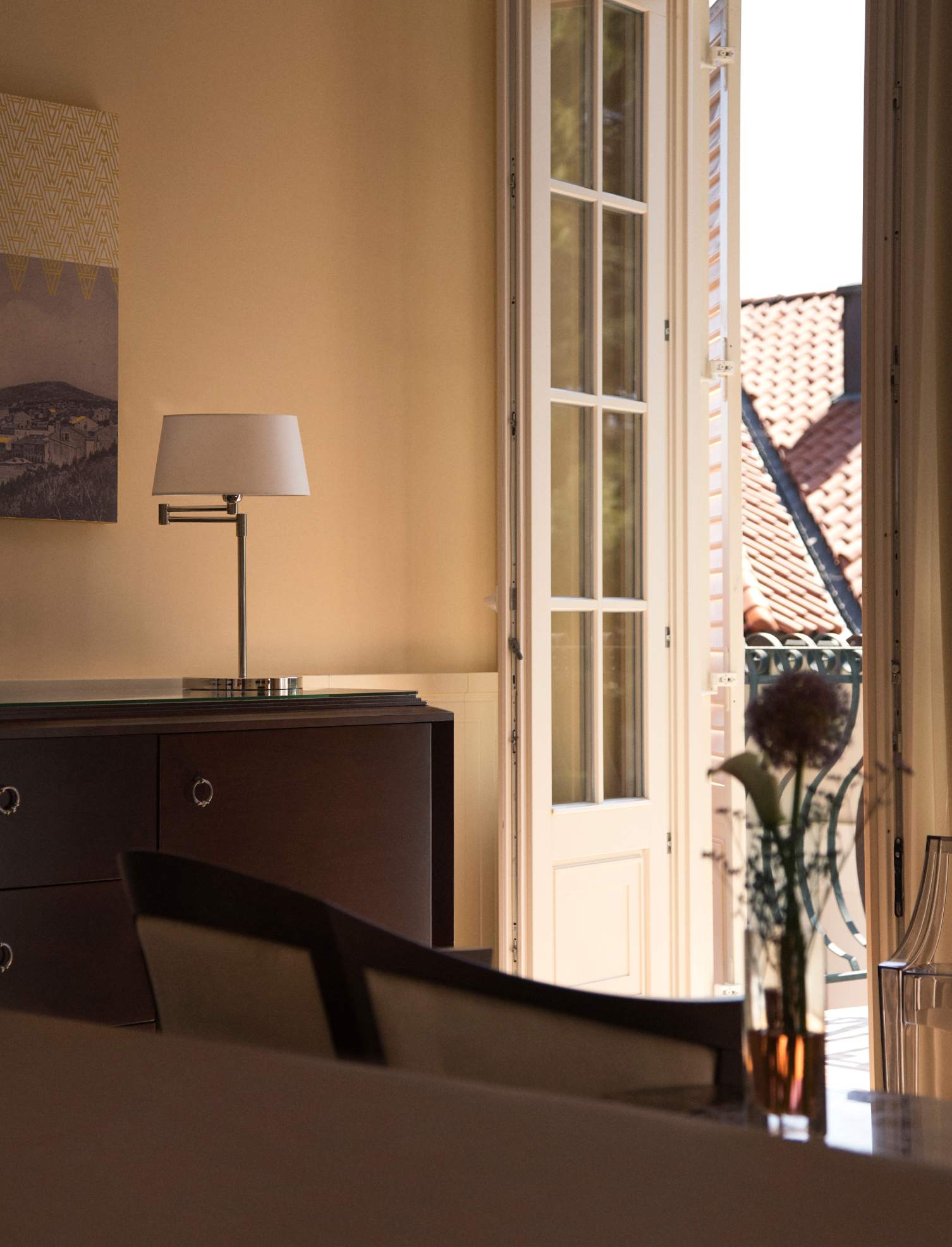 Interior shot with warm lighting, showcasing a sleek table lamp, a dark wood dresser, and open white shutters with a view of red rooftops