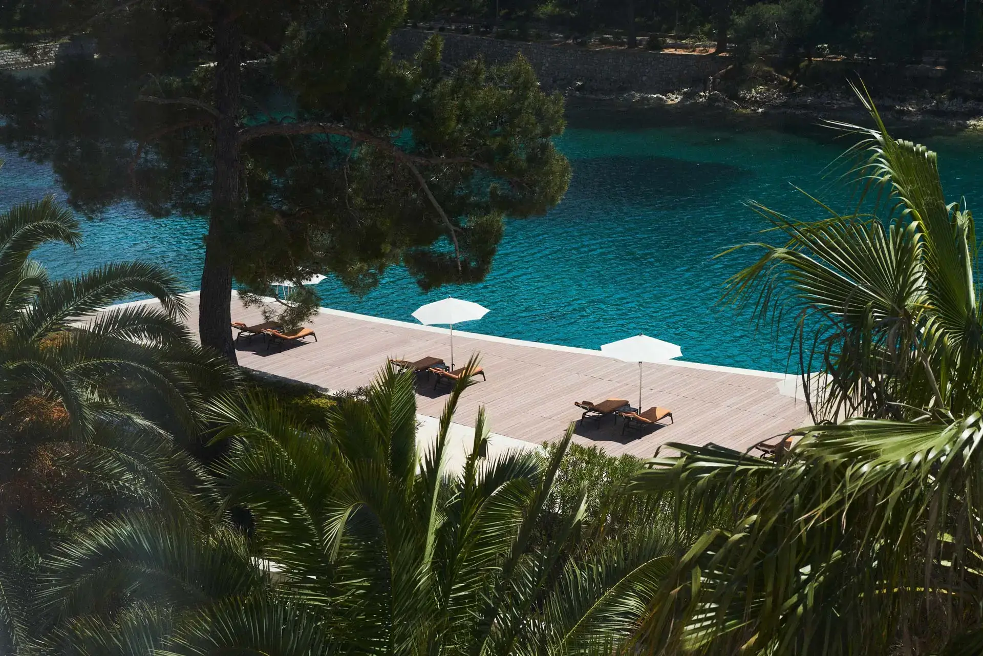 Secluded deck with sun loungers shaded by umbrellas, surrounded by lush palms and overlooking tranquil turquoise waters.