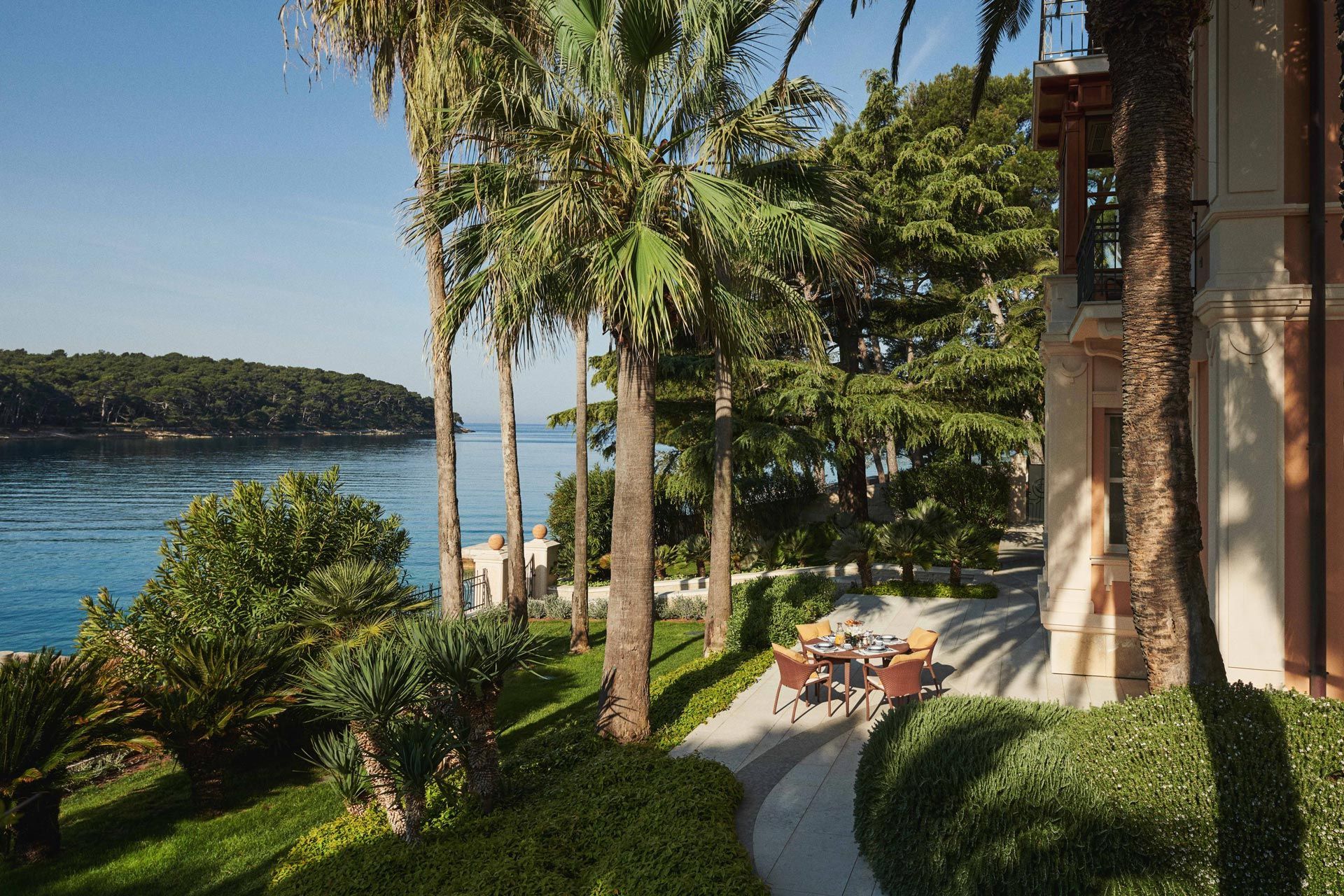 Seaside garden with tall palm trees and a table set for a meal, next to an elegant villa with a view of the bay