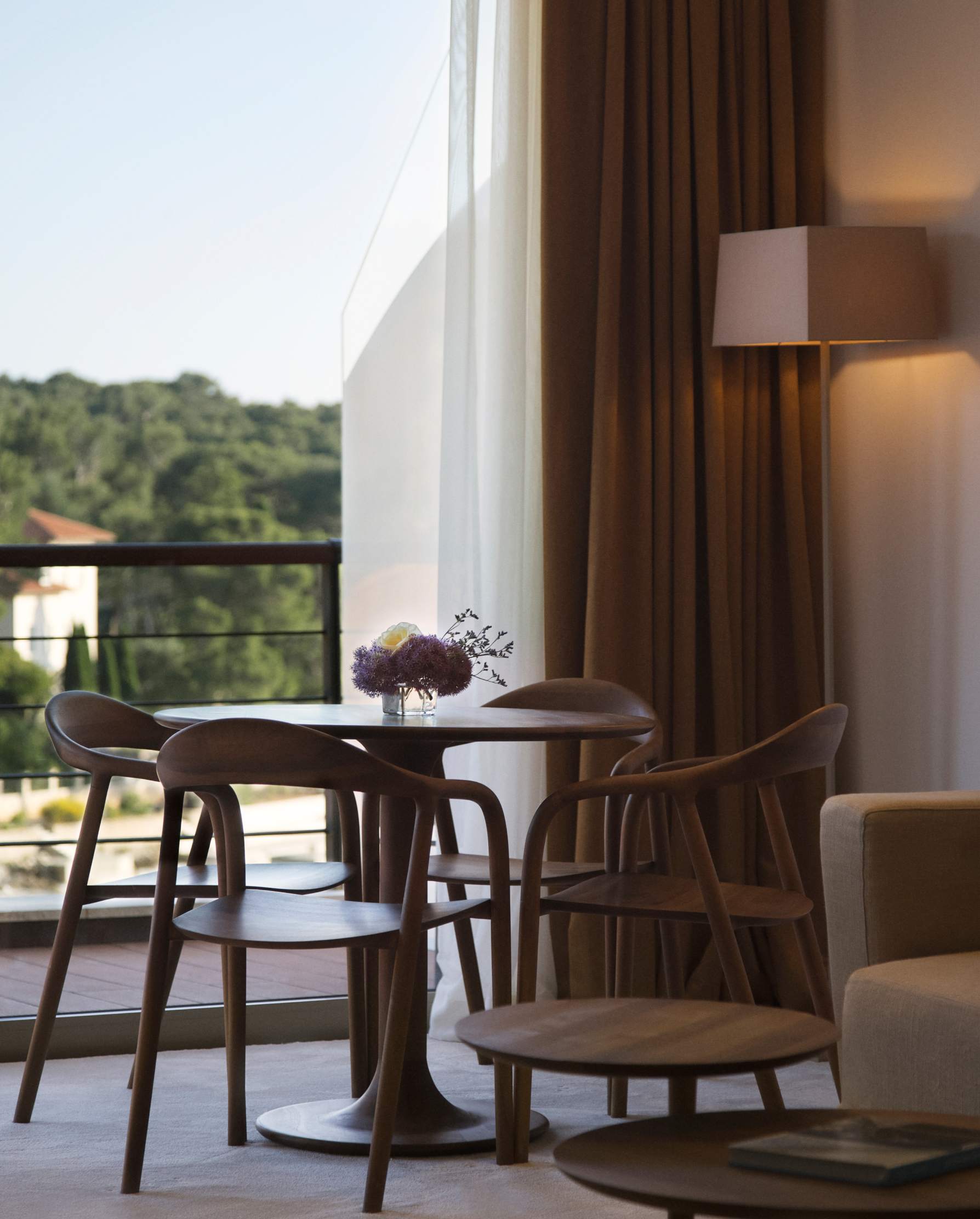 A stylish seating area with a round wooden table and chairs, near a window with sheer curtains offering a view of the outdoor balcony and lush trees