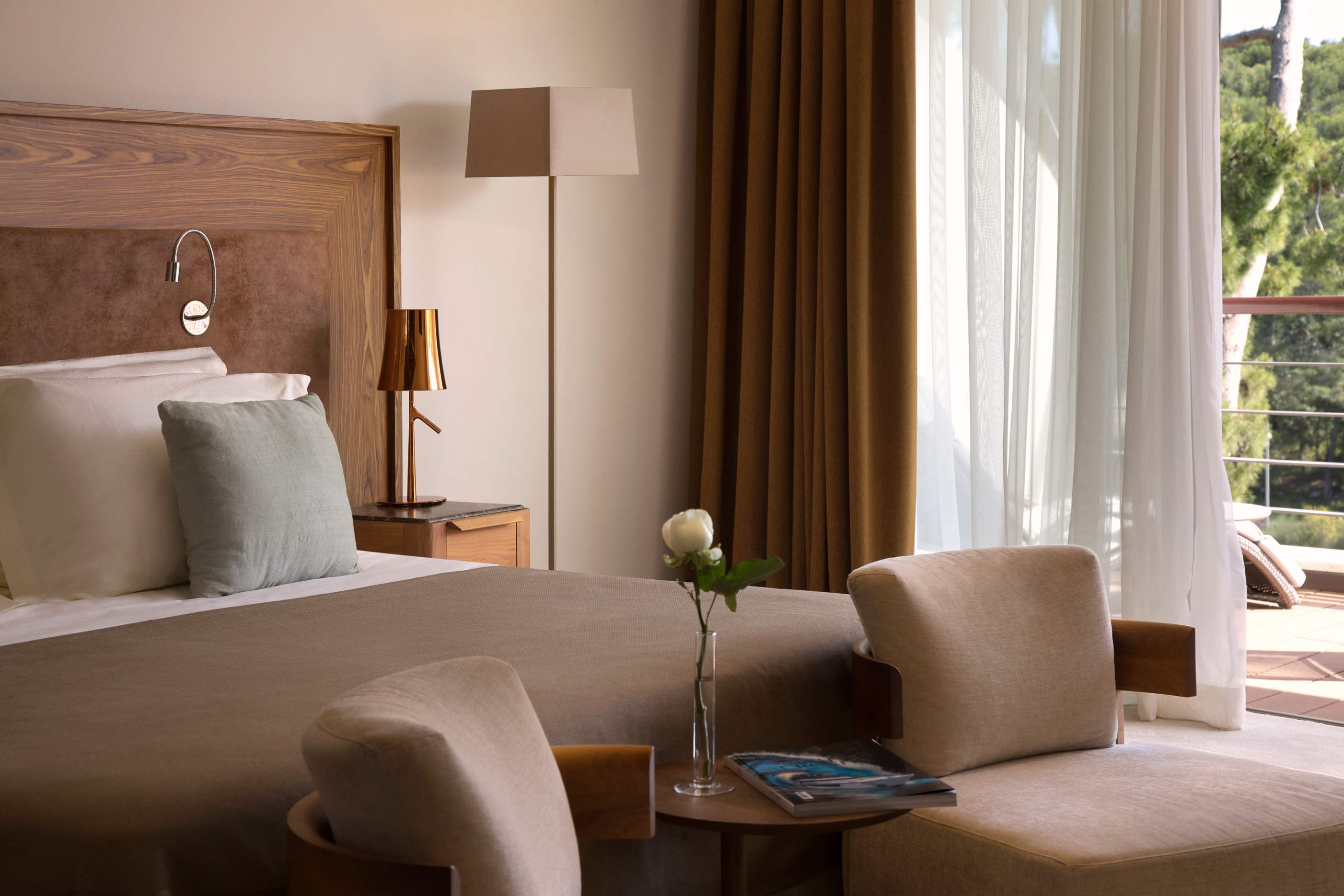 A serene hotel suite with a plush bed, armchairs, and a balcony view, bathed in natural light