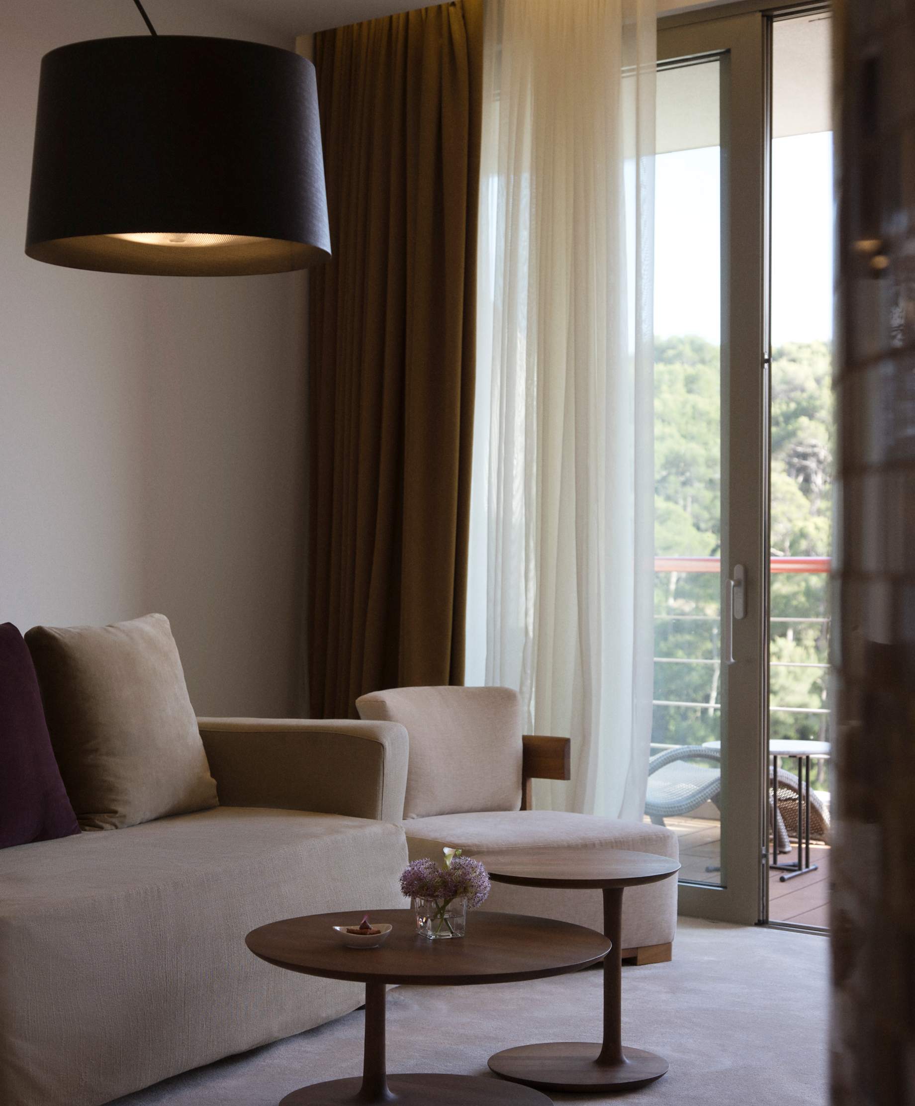 Interior of a hotel room with a beige sofa and armchairs, wooden coffee tables, and sheer curtains, leading to a balcony with a green view.