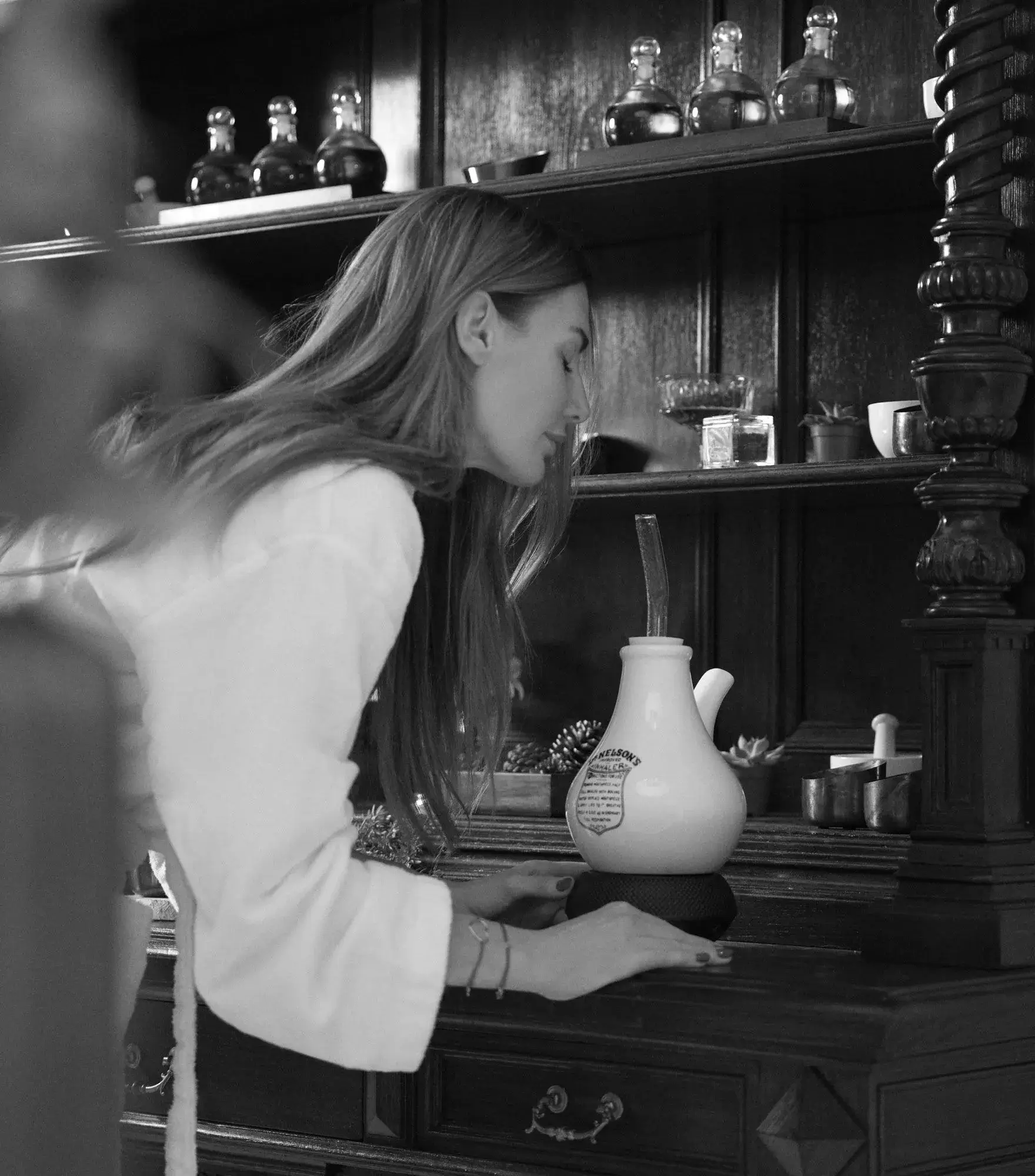 A black and white image of a woman using an inhalation device while standing at a wooden bar in a spa, surrounded by various bottles