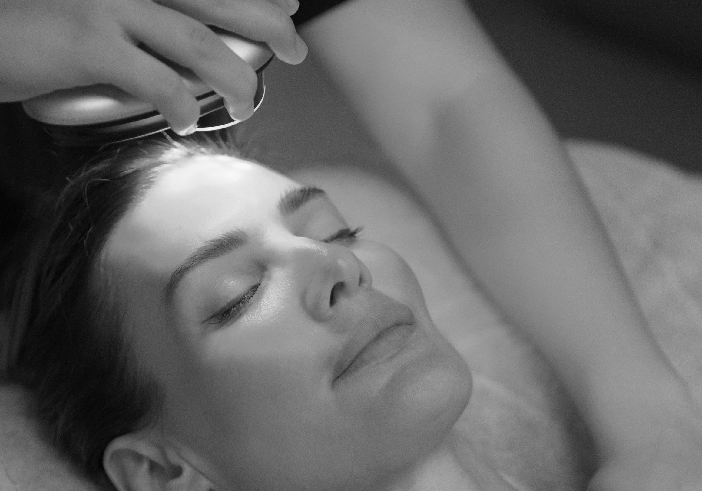 A black and white image of a woman receiving a facial treatment with a red light therapy device