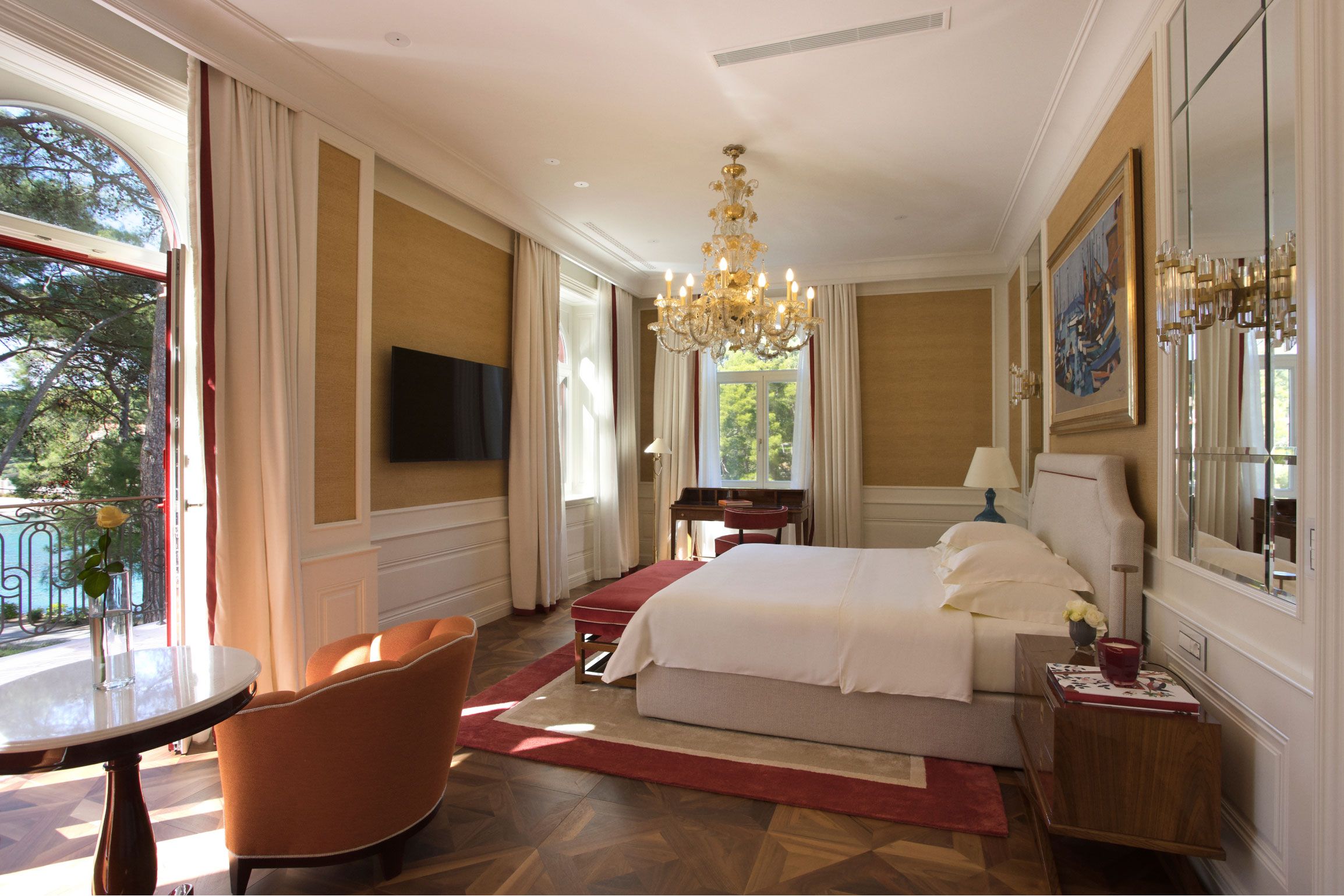 An opulent bedroom featuring a plush bed, an elegant chandelier, a classic wooden desk by a window with a view, and tasteful decor