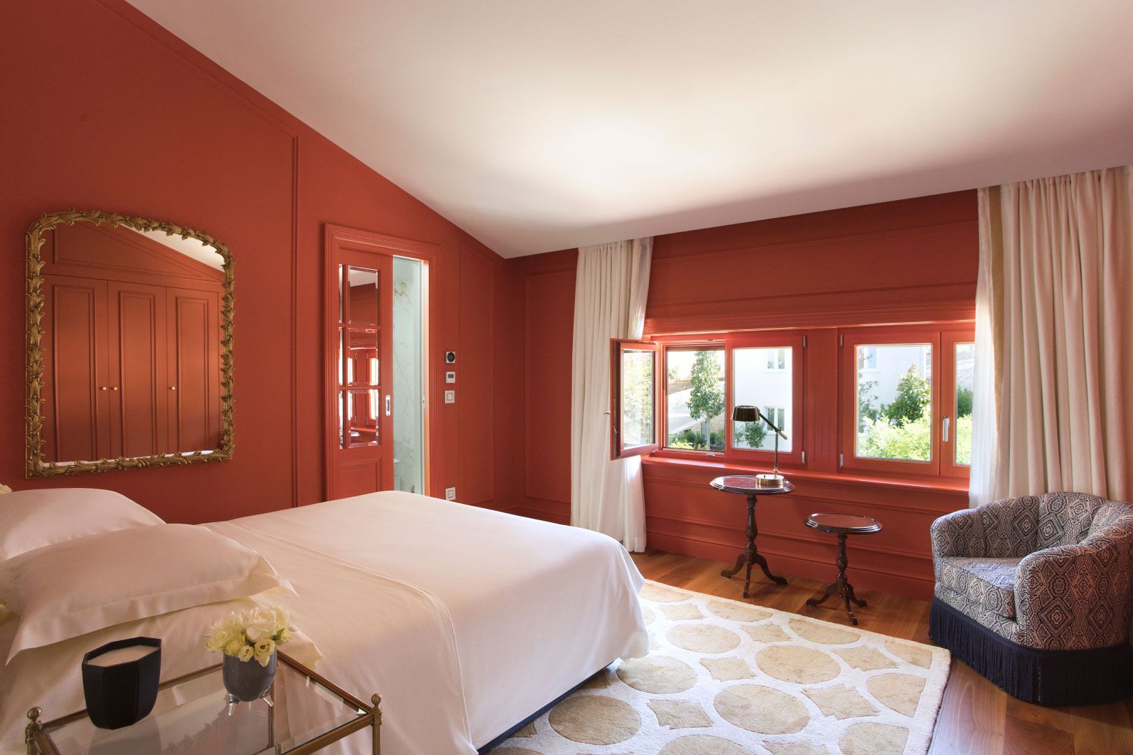 A cozy bedroom with rich red walls, a comfortable bed, traditional furnishings, and a view of the outdoors through bright windows