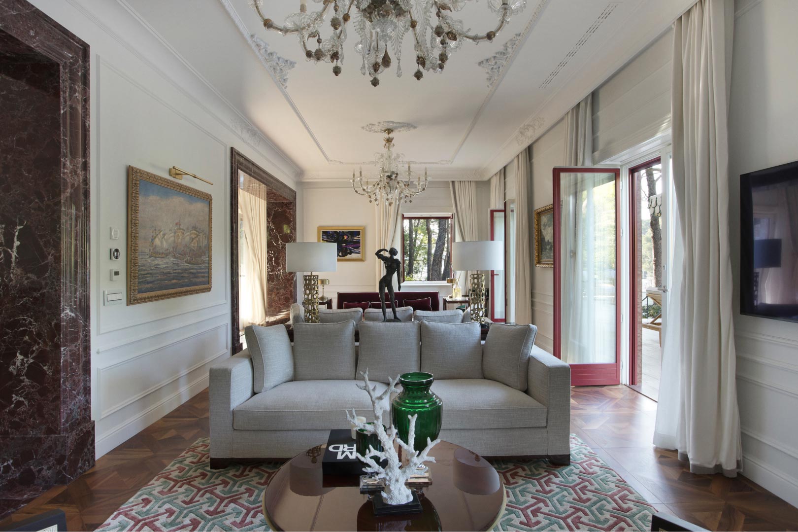 A sophisticated living room with a plush sofa, intricate area rug, ornate chandeliers, and framed artwork, complemented by marble columns and elegant drapery