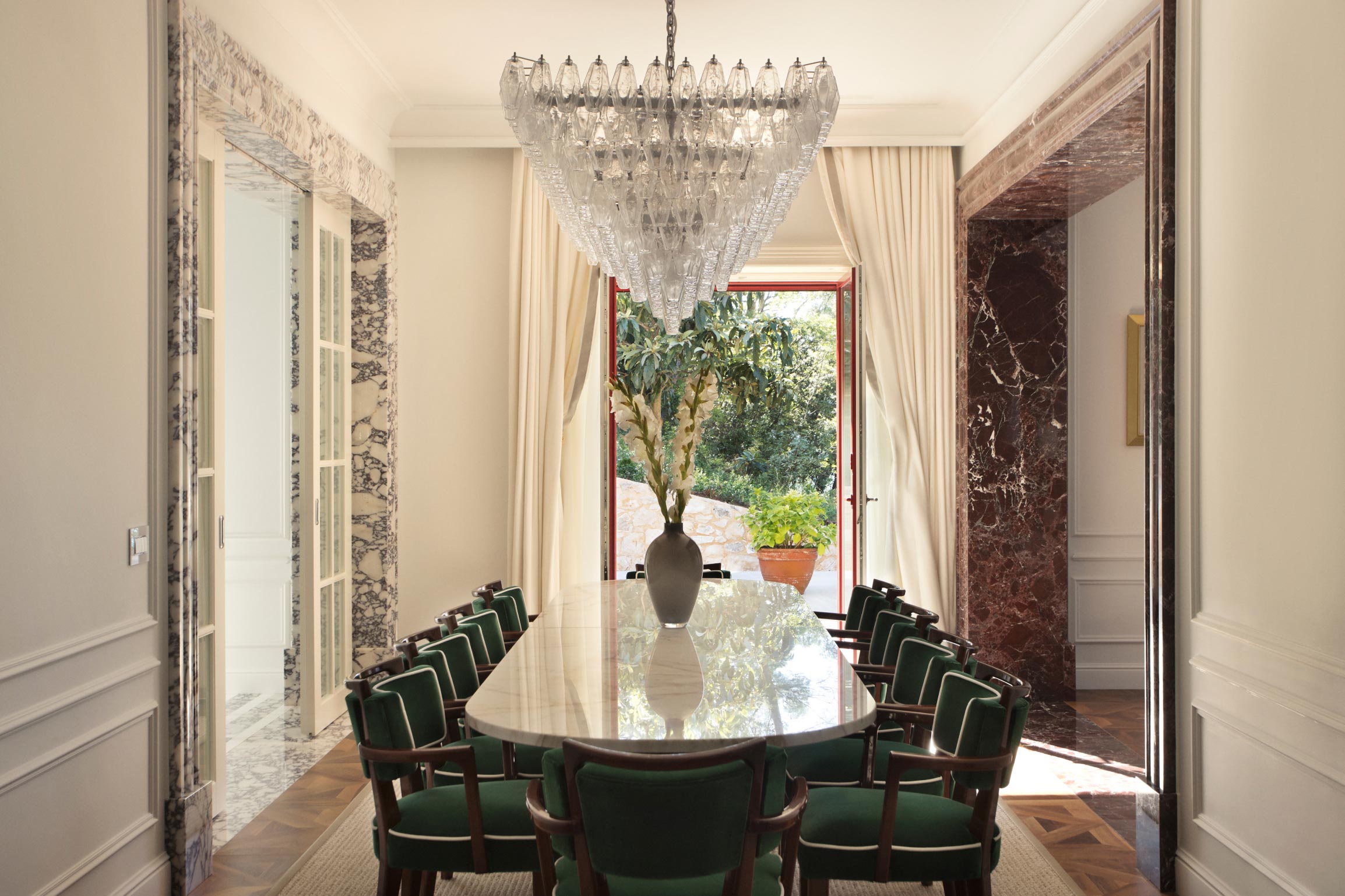 An elegant dining area with a long table, green upholstered chairs, and a striking glass chandelier, offering a view to a verdant outdoor space