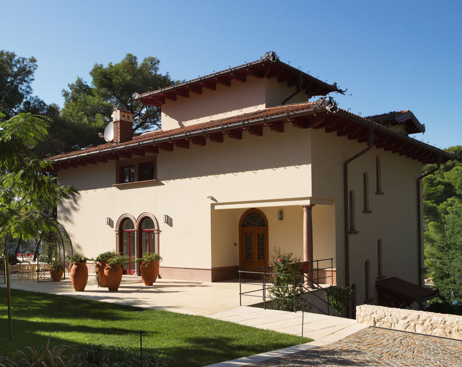 A traditional villa with a terracotta roof, surrounded by lush greenery, featuring arched windows and large terracotta planters, nestled in a tranquil natural setting