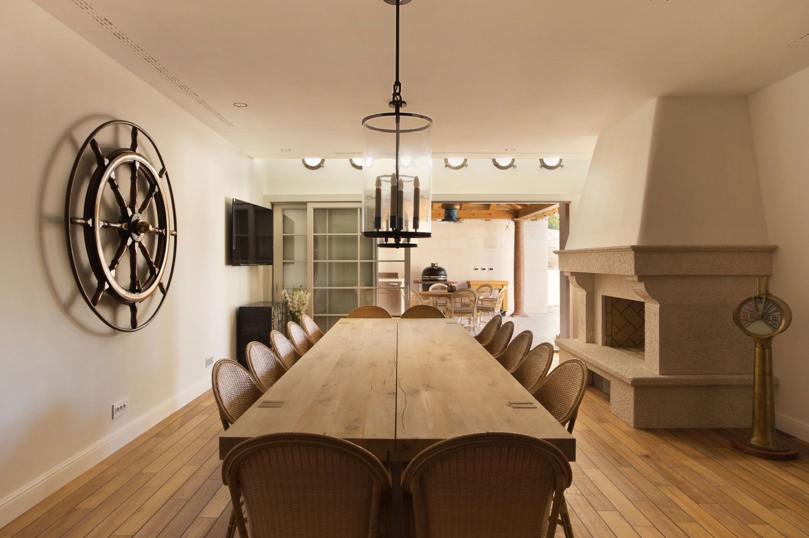 A spacious dining area with a long wooden table, cane-back chairs, and a large decorative wheel on the wall, complemented by a classic fireplace and a view to an outdoor patio