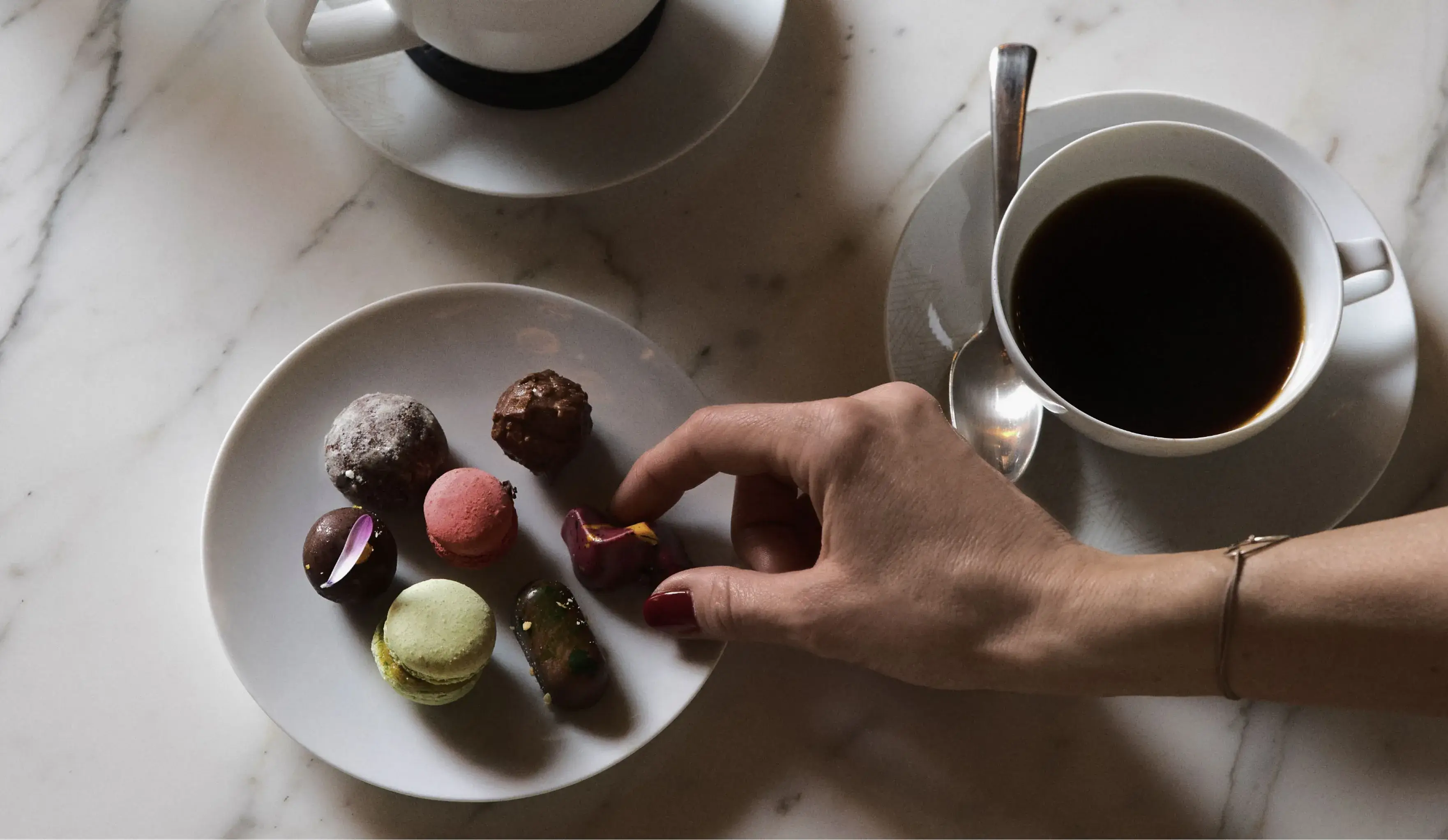 A hand selecting a treat from a plate of macarons next to a cup of coffee and a creamer on a marble table