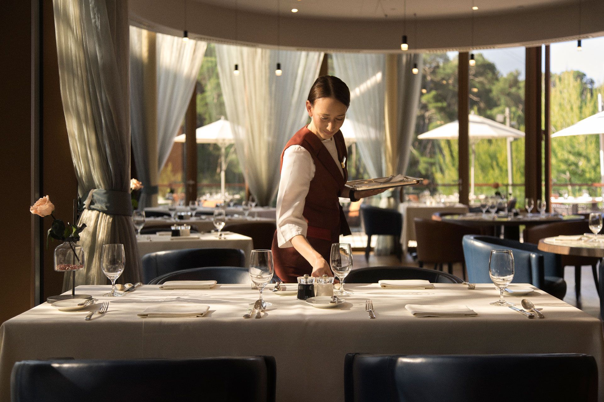  A waitress setting a table in an elegant dining room with large windows and soft natural light