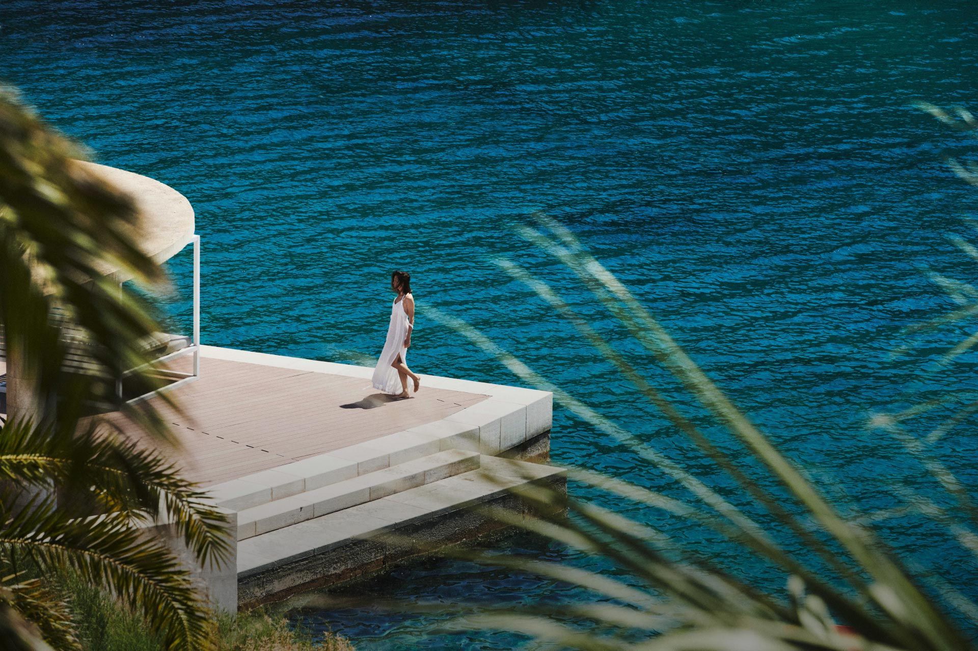 A woman in a white dress walking on a seaside deck, with the deep blue sea in the background.