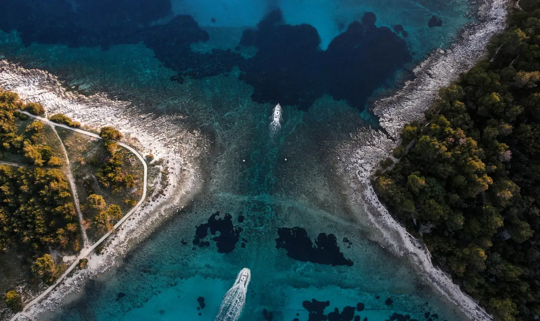 A bird's-eye view of a coastal scene where two boats are moving through clear blue waters near a rocky shore, lined with trees