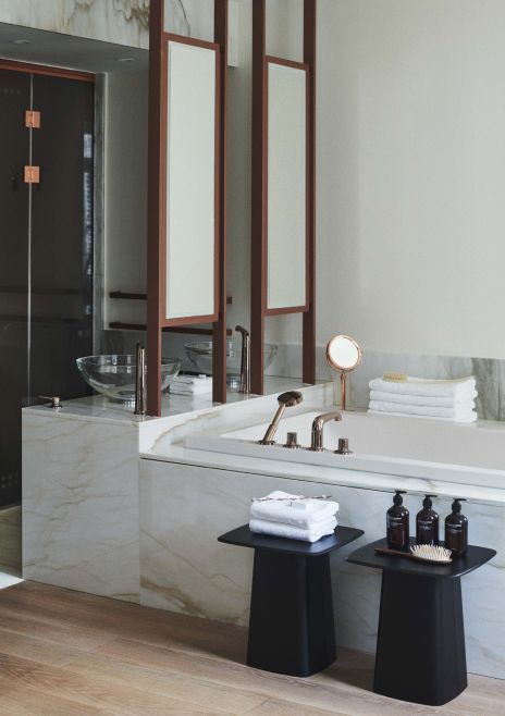 A modern bathroom with marble walls and countertops, featuring sleek mirrors, a glass bowl sink, and minimalist black accent tables with toiletries.