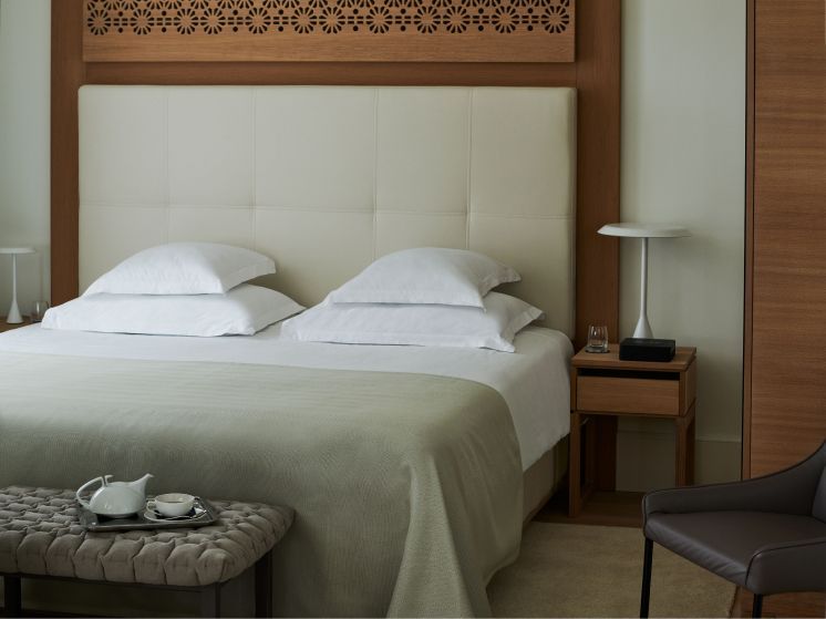 A neatly made bed with a padded headboard, flanked by a nightstand and lamp, with a subtle earth-toned color palette creating a calm bedroom atmosphere.