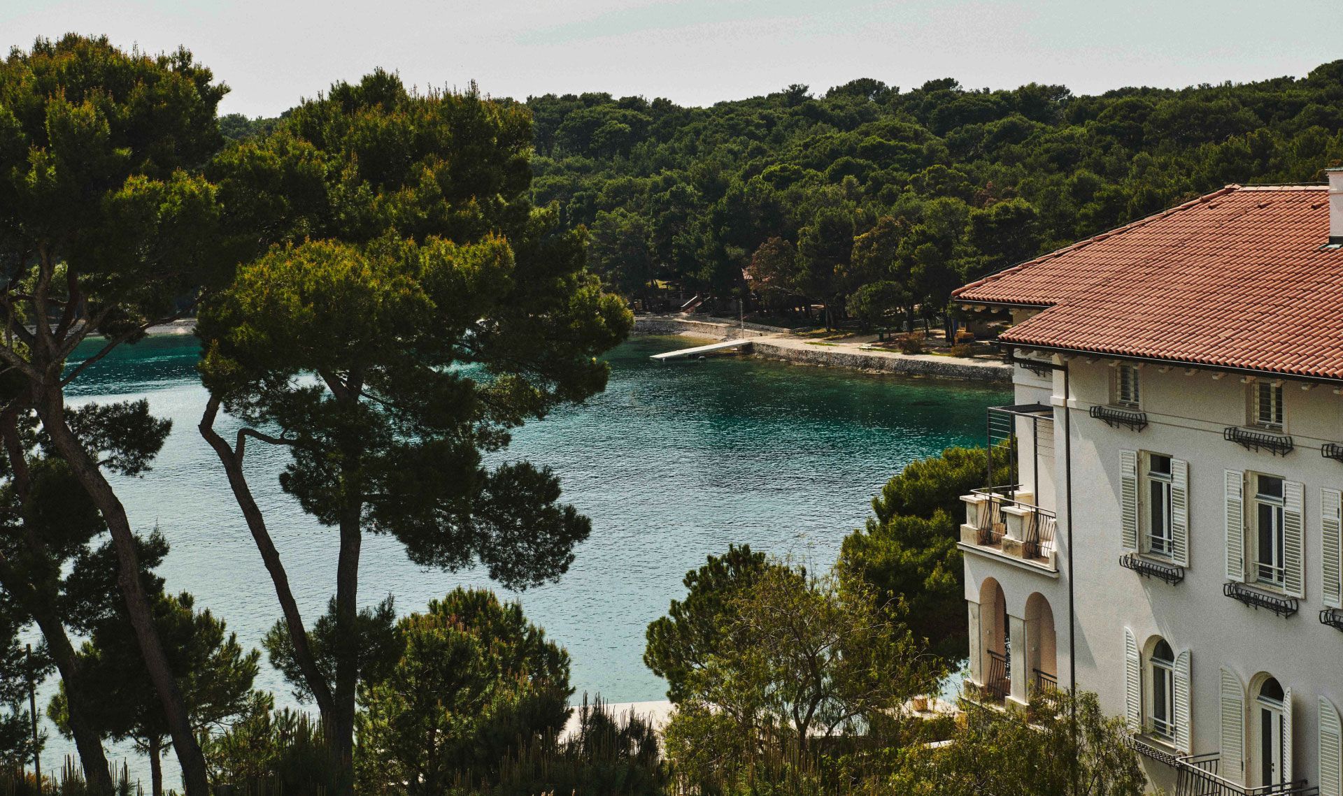 Scenic view of a seaside villa with balconies overlooking a tranquil bay surrounded by lush pine trees