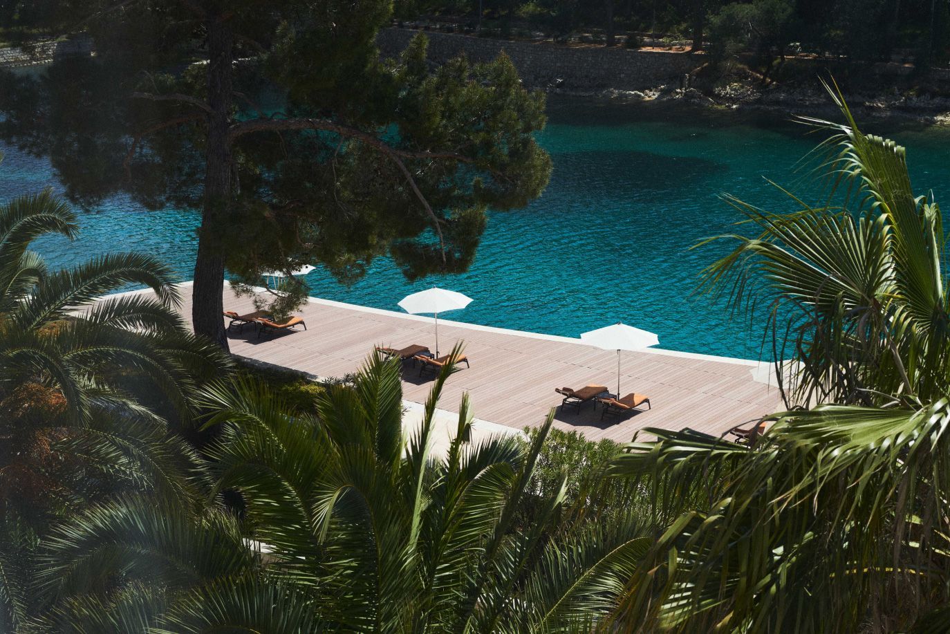 Secluded deck with sun loungers shaded by umbrellas, surrounded by lush palms and overlooking tranquil turquoise waters.