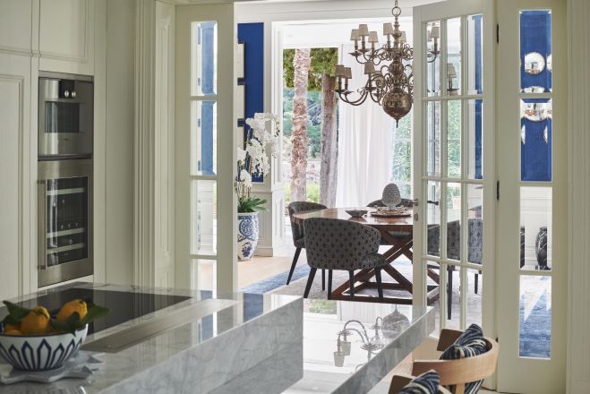 A view from a marble kitchen countertop through open French doors to a dining area with elegant furnishings and a classic chandelier, embodying a bright and sophisticated interior design.