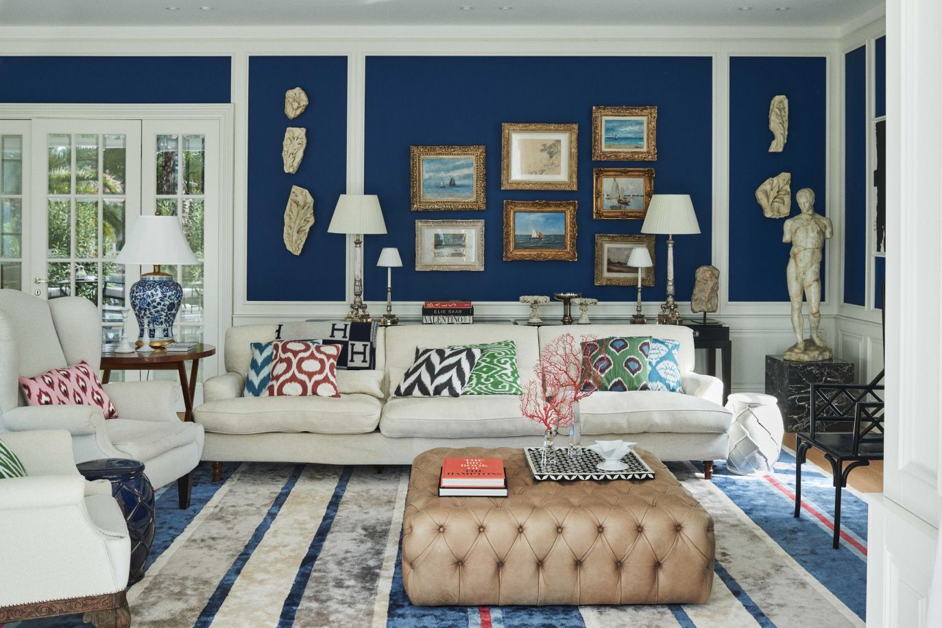 A luxurious living room with bold blue walls, adorned with an array of framed artwork, complemented by a mix of patterned and solid cushions on plush seating, and a classical statue adding a touch of elegance.