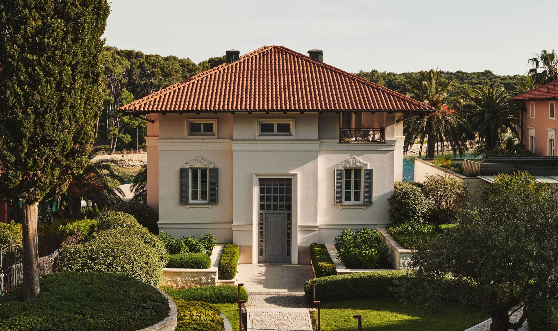 A well-manicured villa with a tiled roof, flanked by lush greenery and overlooking a beach with palm trees.