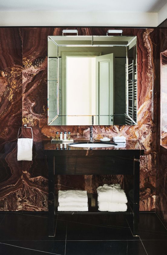 An elegant bathroom vanity with a large mirror set against a striking marble wall in warm brown and red tones, complemented by black glossy countertops and neatly folded white towels.