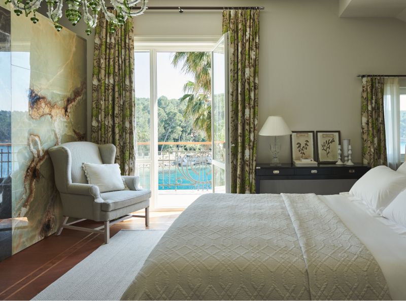 A tranquil bedroom with neutral tones, an armchair by the window, offering a view of palm trees and a waterfront balcony, accentuated with floral curtains and a unique marble feature wall.