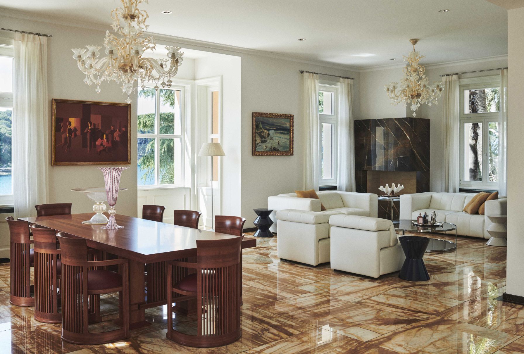 An elegant dining and living space with marble flooring, featuring a wooden dining table with chairs, ornate chandeliers, plush white sofas, and art pieces complementing the room's luxurious aesthetic.