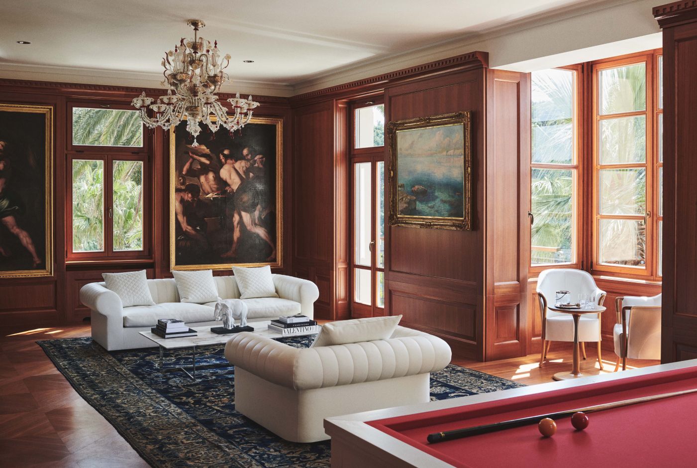 Classic living room with a white sofa, billiards table, ornate chandelier, and framed artwork, with sunlight streaming in through window shutters.