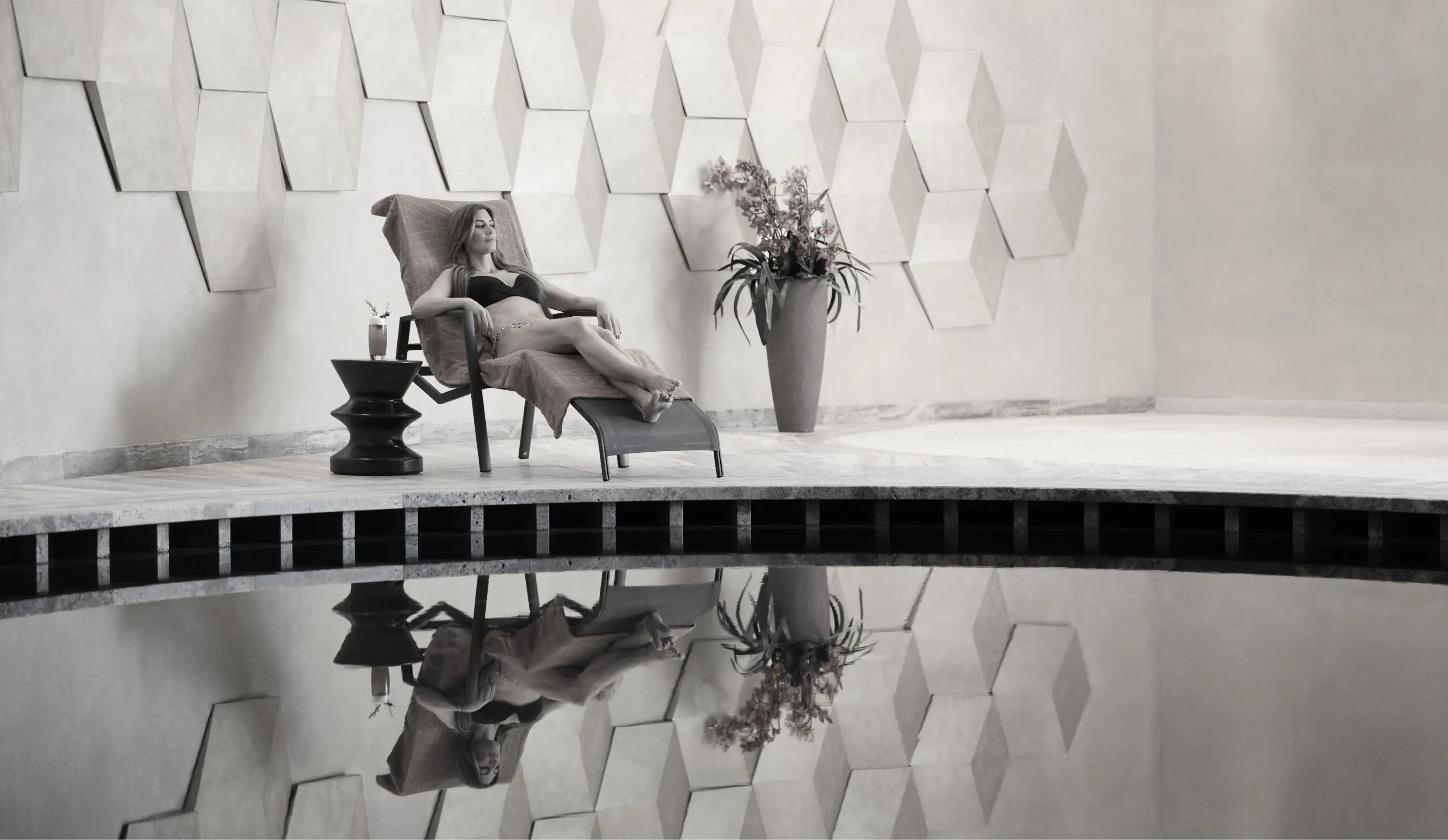 A black and white image of a relaxed woman lounging on a chair by a reflective pool, with a drink on a side table and geometric wall art in the background