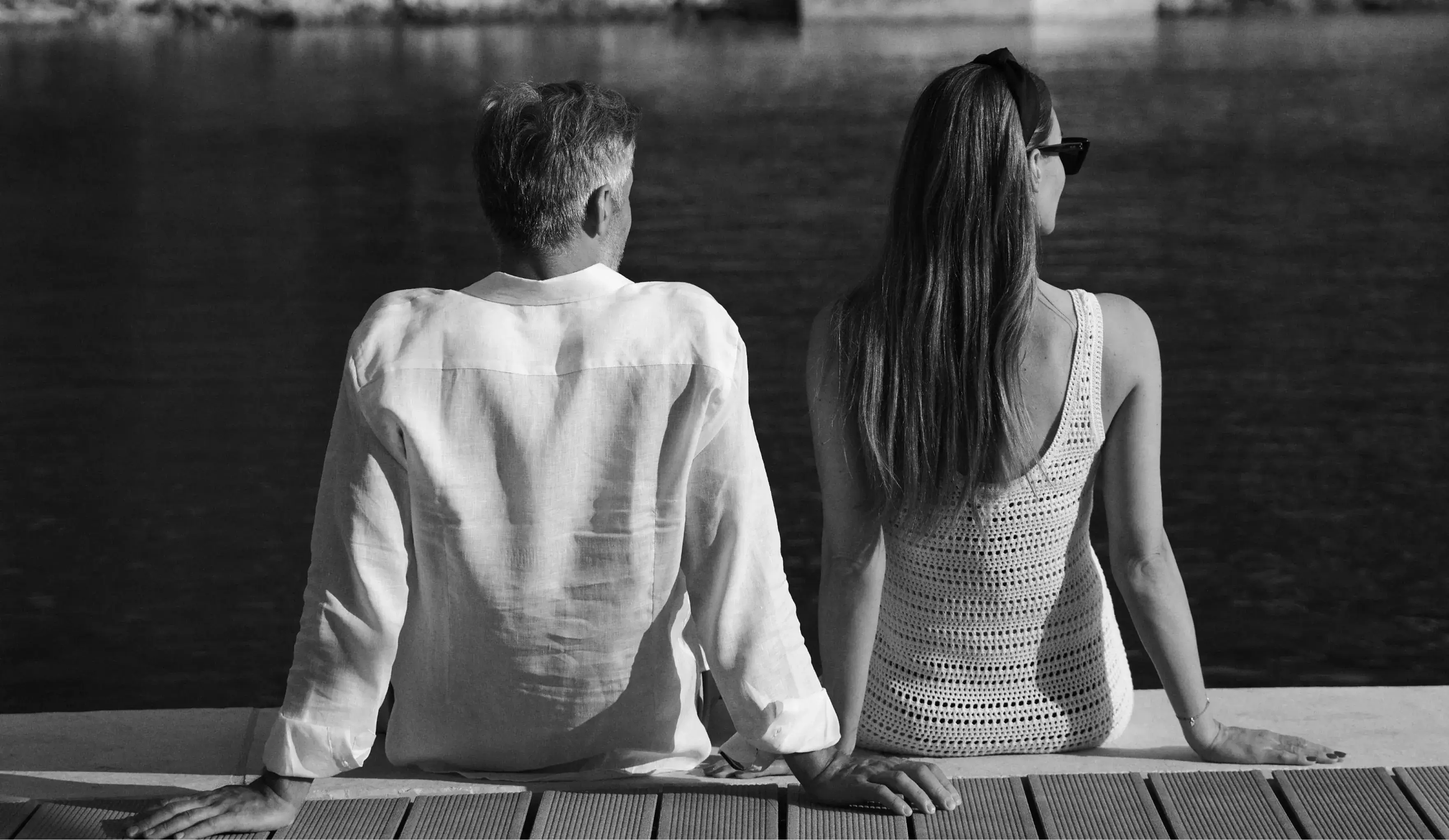 A black and white image of a couple sitting by the water's edge, looking out across the bay