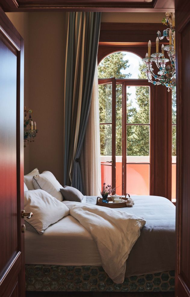 A view of a cozy bedroom with an inviting bed, elegant drapes, a colorful chandelier, and a sunny window overlooking trees