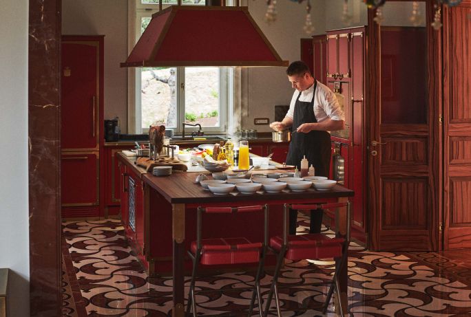 A chef preparing a meal in a luxurious kitchen with red wooden cabinetry, a matching island, and elegant patterned flooring