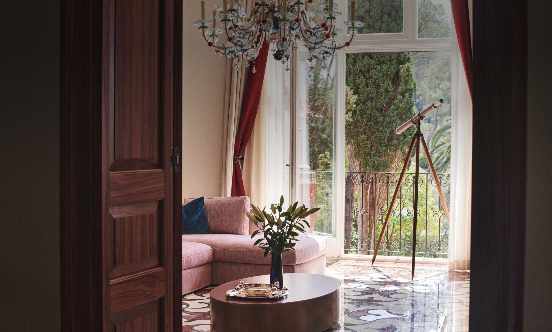 A plush pink sofa and ornate chandelier in a sunlit room, with a balcony door open to a garden view and a vintage telescope