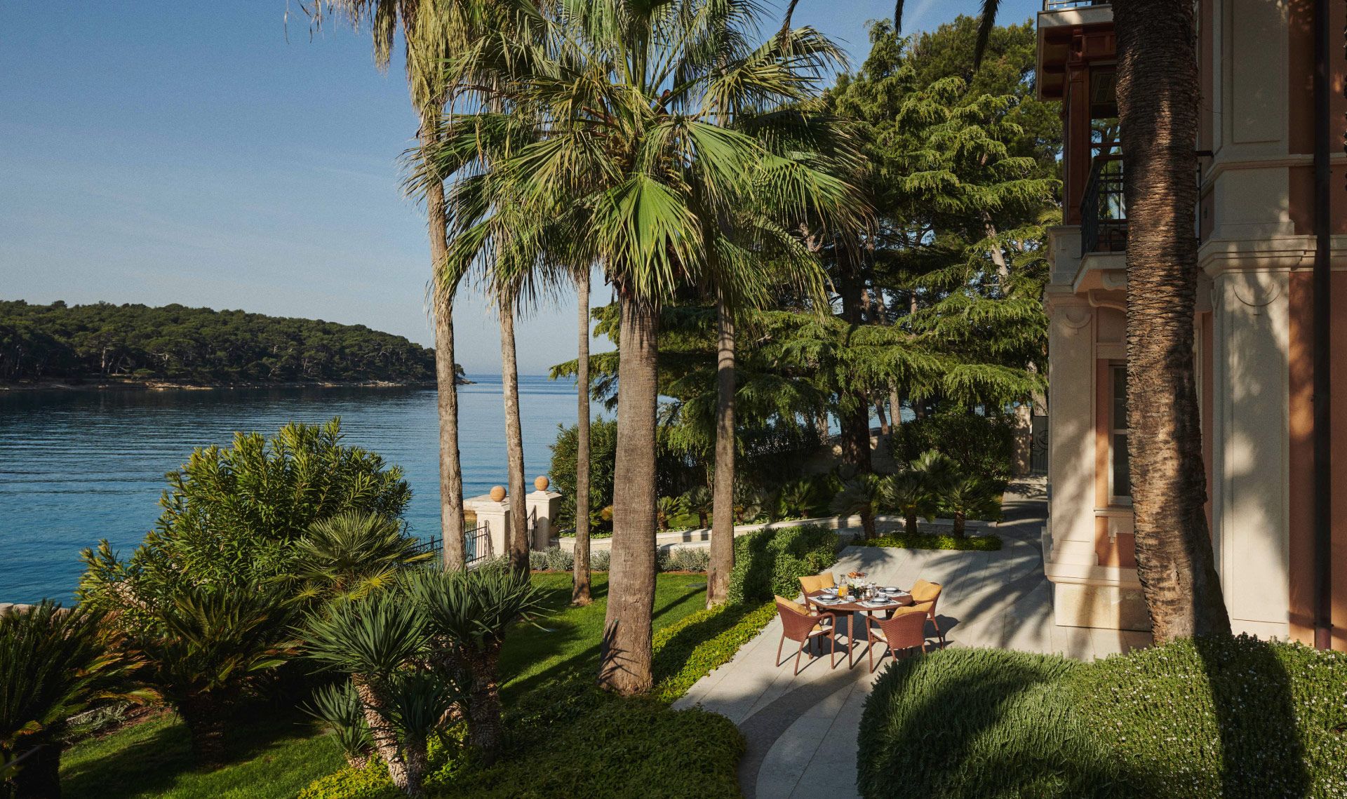 Seaside garden with tall palm trees and a table set for a meal, next to an elegant villa with a view of the bay