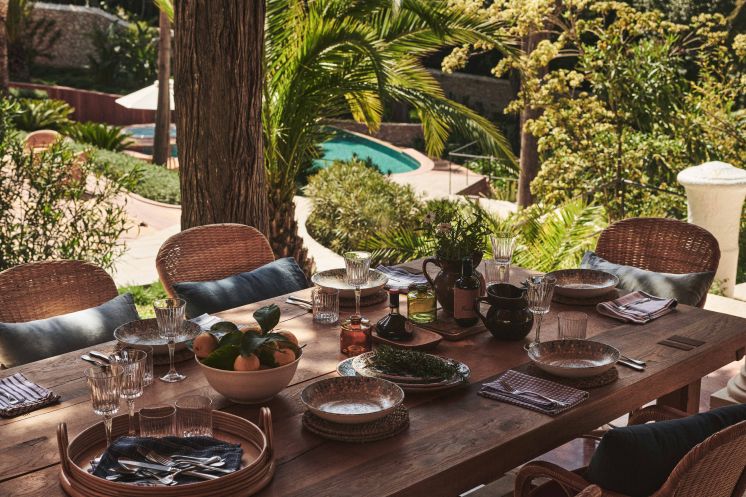 An outdoor dining table set with wicker chairs on a terrace, overlooking a pool amidst lush palm trees, ready for a meal in a serene setting.