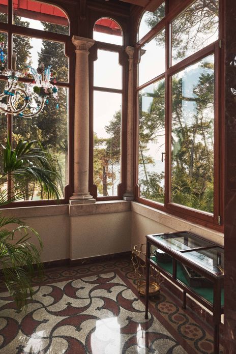 A bright sunroom with tall arched windows and a decorative tiled floor, offering a scenic view of a forest and water through the foliage.