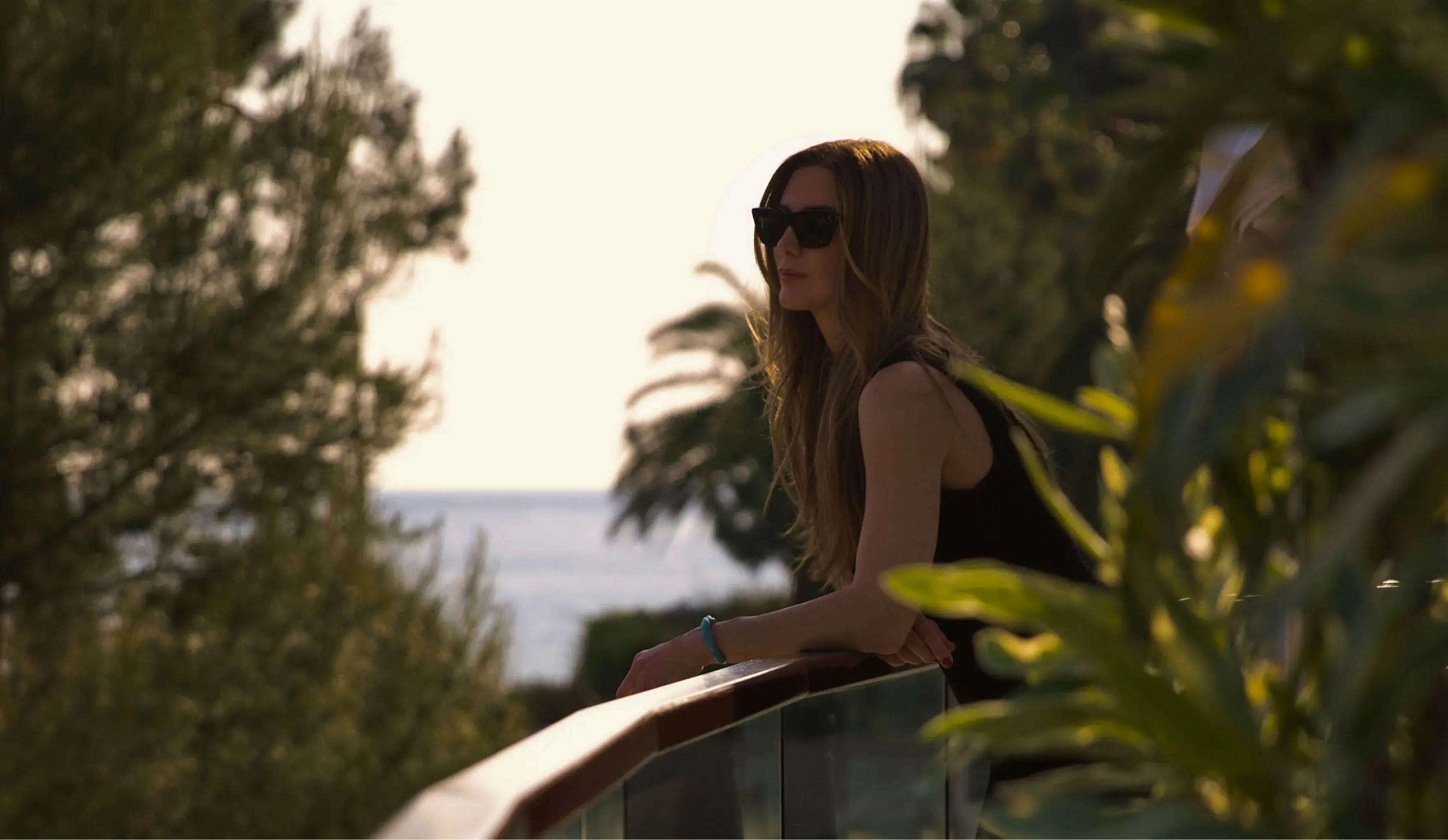 A woman in a casual outfit leaning on a glass railing, surrounded by lush greenery, with a glimpse of the sea in the background