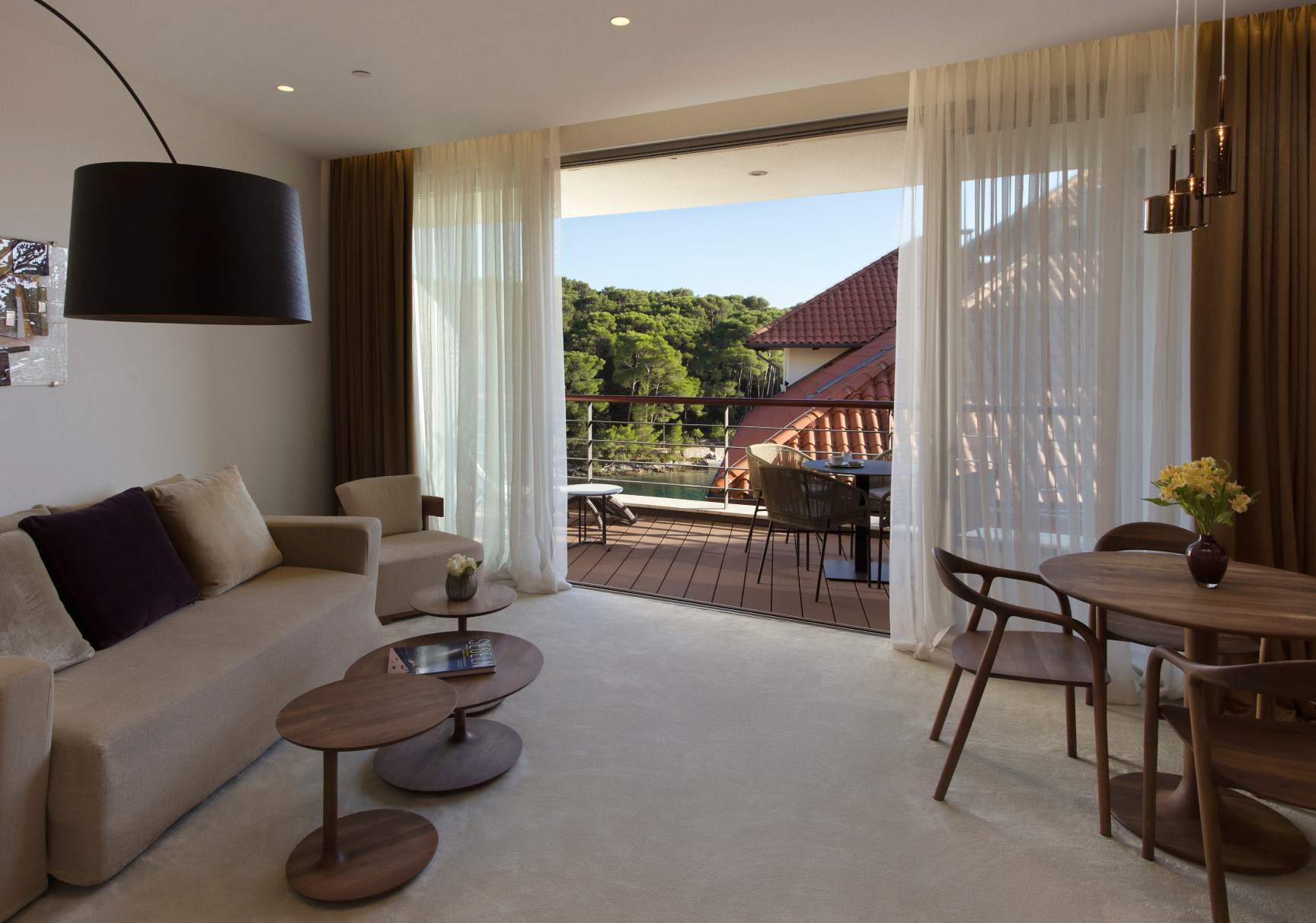 A spacious and modern living area with a beige sofa, wooden tables, and large windows leading to a balcony with an outdoor dining set and a view of lush greenery