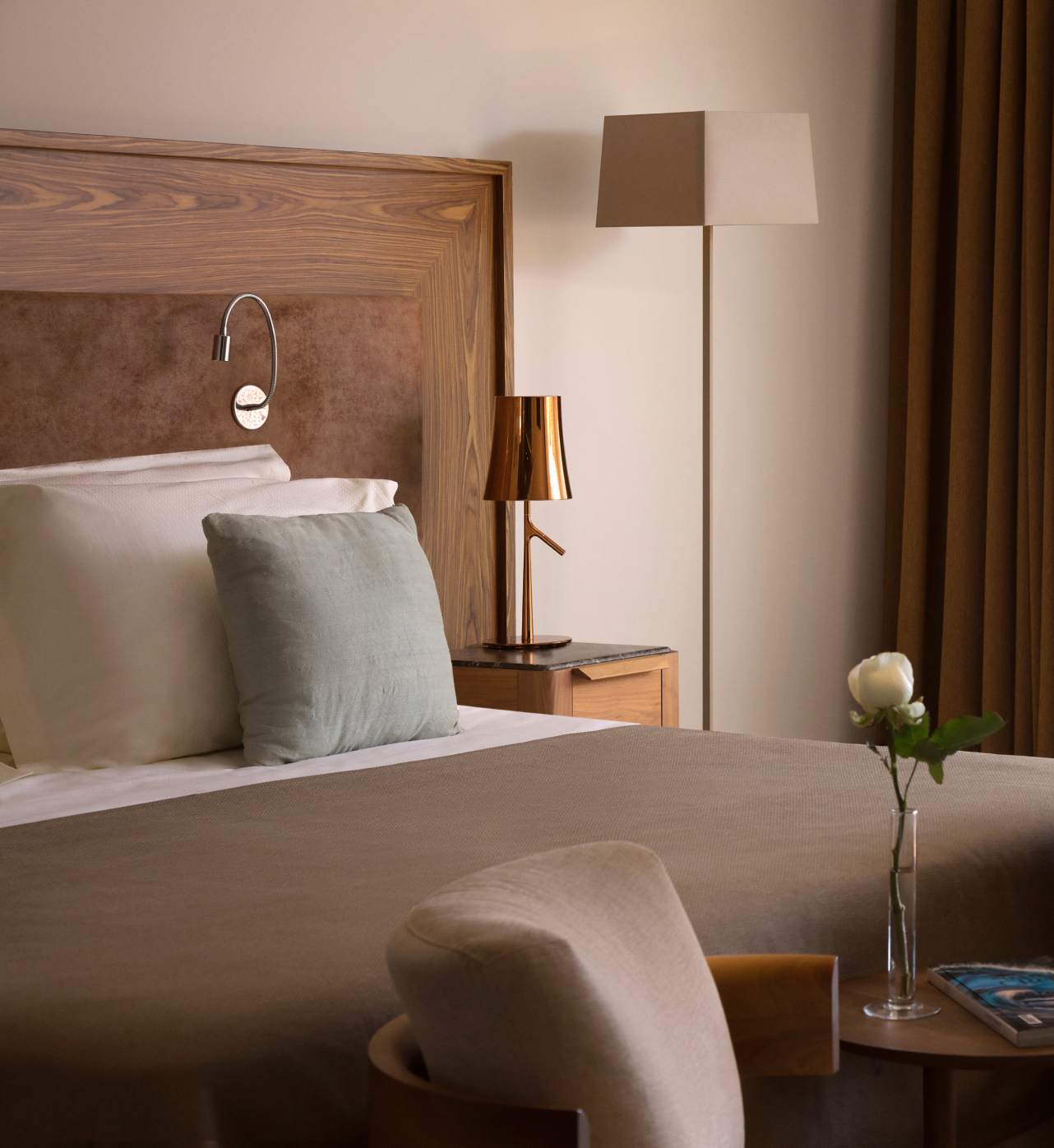 A warm and inviting bedroom corner with a wooden headboard, elegant lamp, and a single white rose in a slender vase on a bedside table