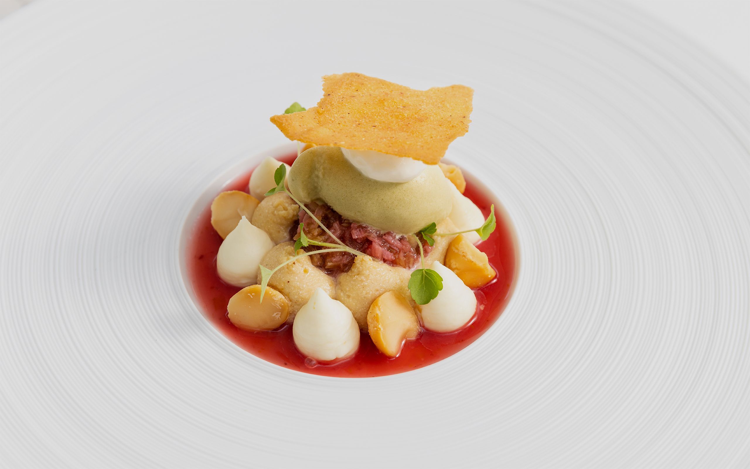 A beautifully plated dessert featuring a vibrant orange sorbet garnished with red berry flakes, fresh raspberries, and delicate cream dollops