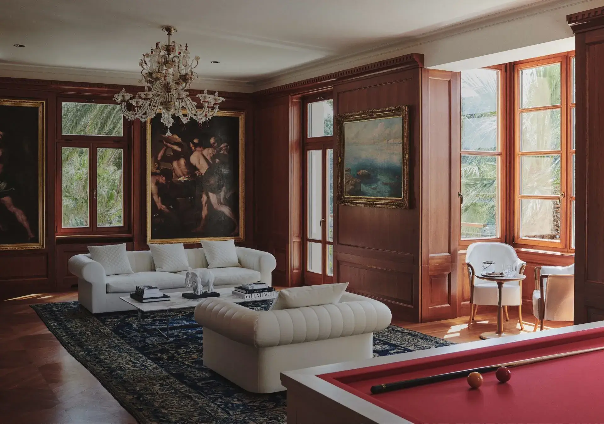 Classic living room with a white sofa, billiards table, ornate chandelier, and framed artwork, with sunlight streaming in through window shutters.